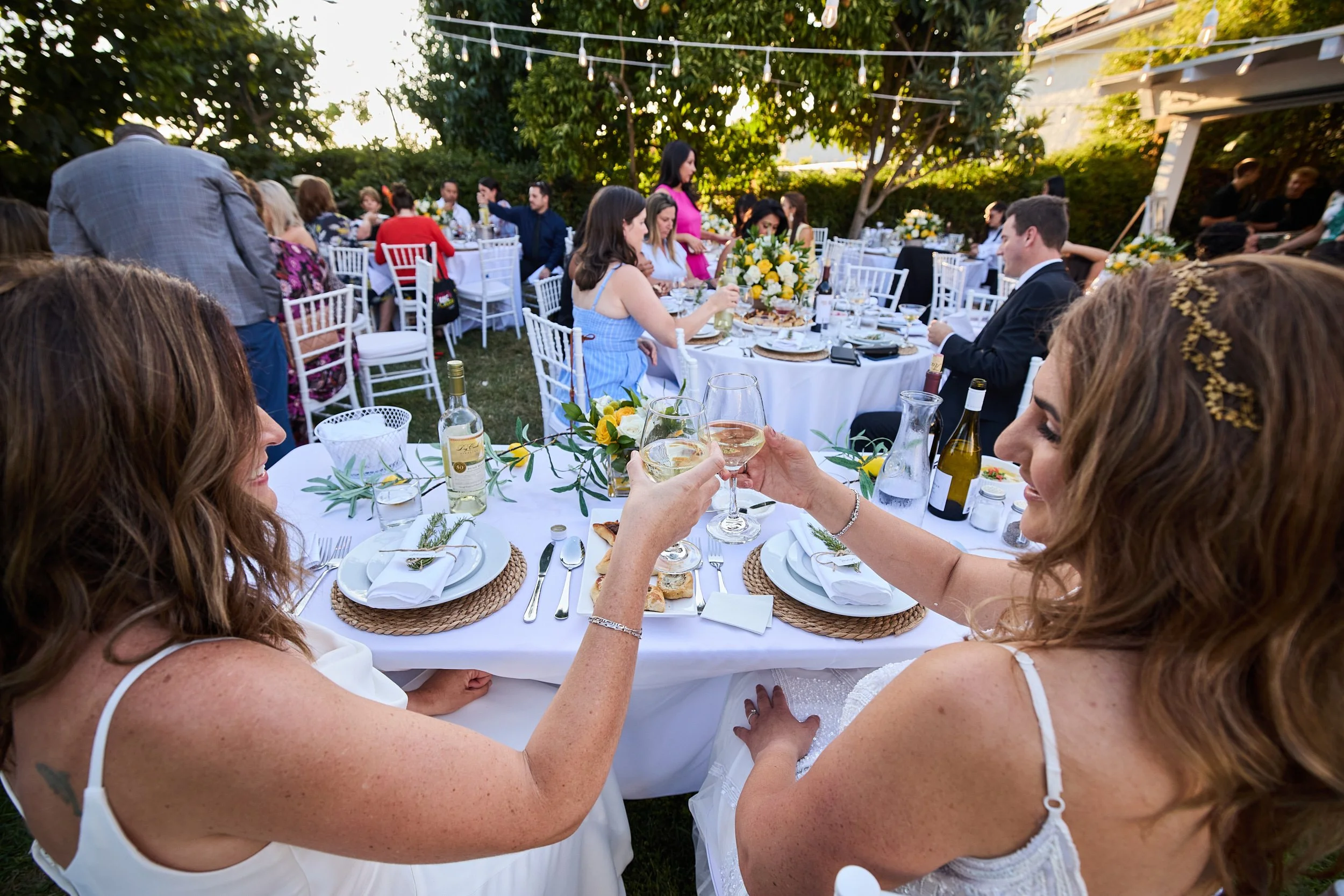 People celebrating at a wedding reception outdoors, toasting with wine glasses, with decorated tables, white chairs, and string lights overhead.