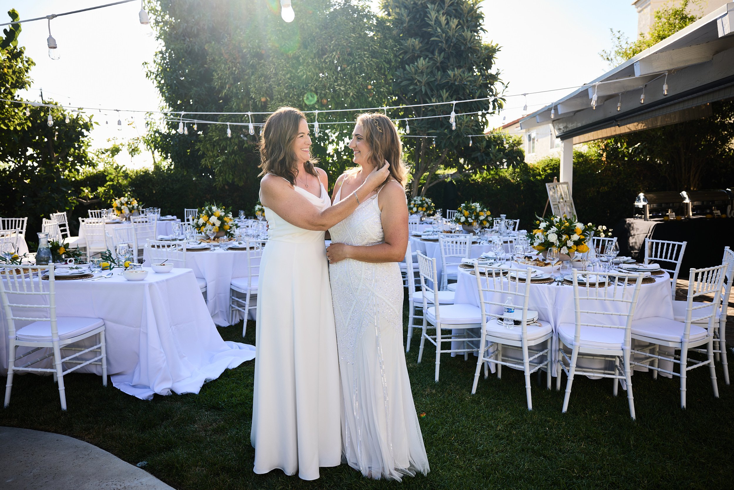 Two women in white dresses sharing a moment in an outdoor wedding reception with decorated tables and chairs, string lights, and trees in the background.