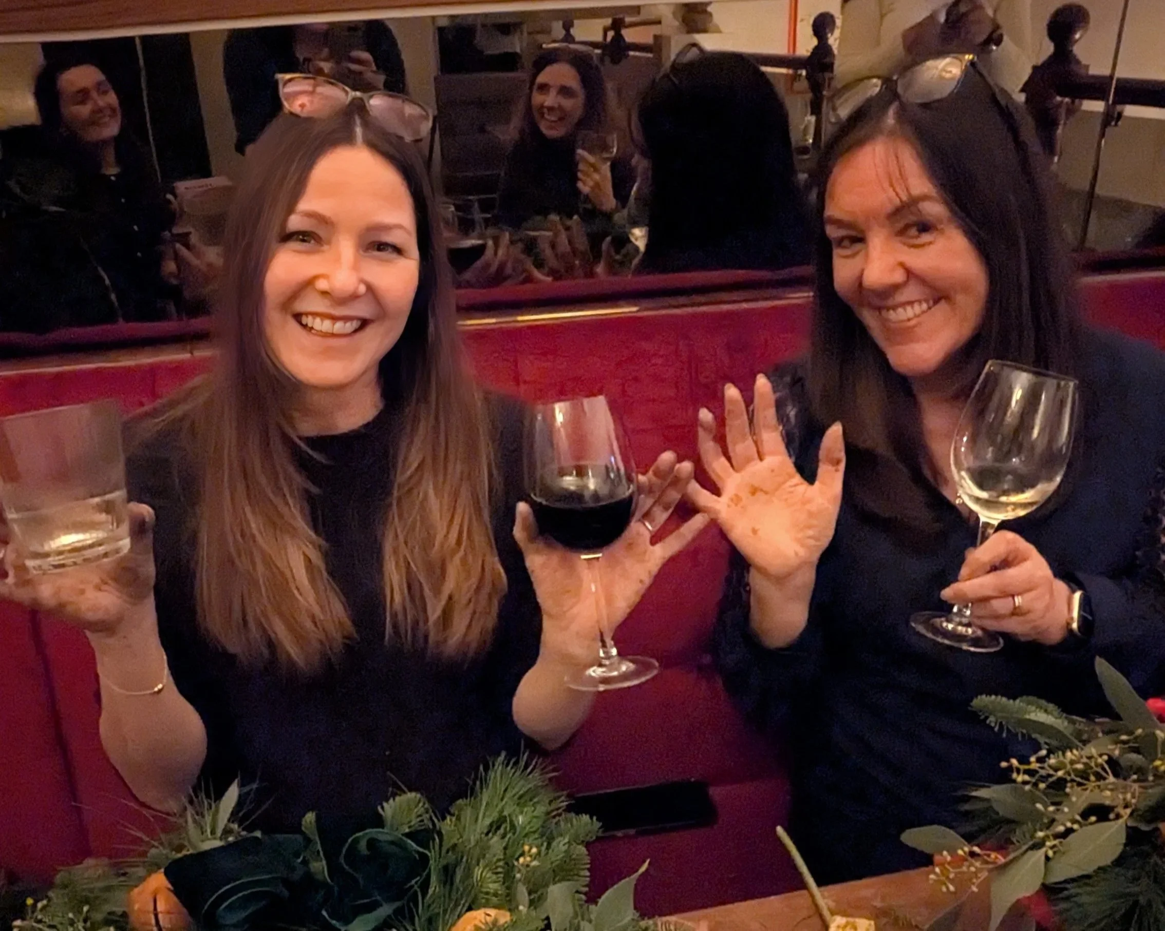 Two women smiling and holding glasses of red wine at a social gathering, with other people and decorations visible in the background.