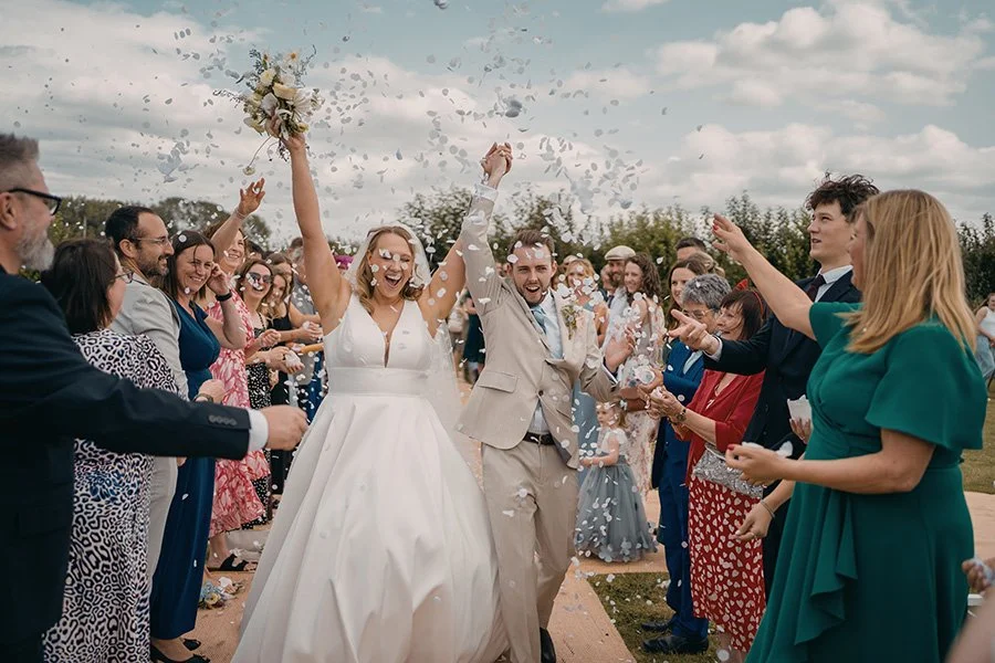 Bride and groom celebrating as they walk through a crowd of guests throwing confetti at an outdoor wedding.