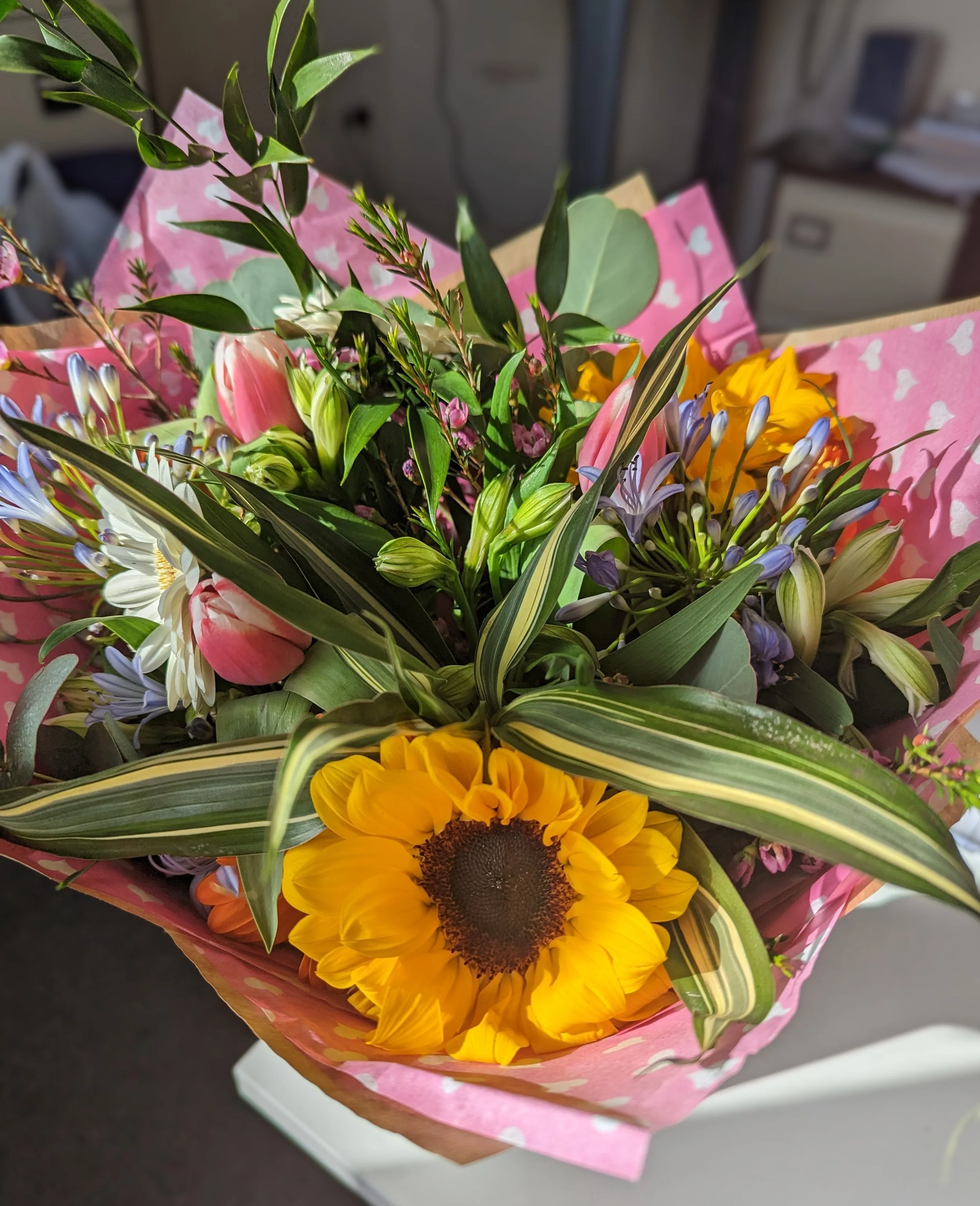 Colorful bouquet of flowers including a large sunflower, lilies, pink tulips, and assorted greenery wrapped in pink paper with white heart patterns.
