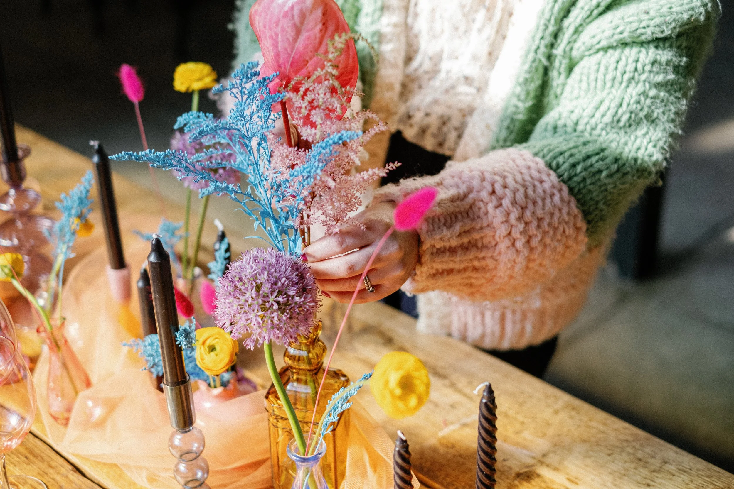 Person arranging colorful flowers in glass vases on a wooden table, with candles and decorative cloth.