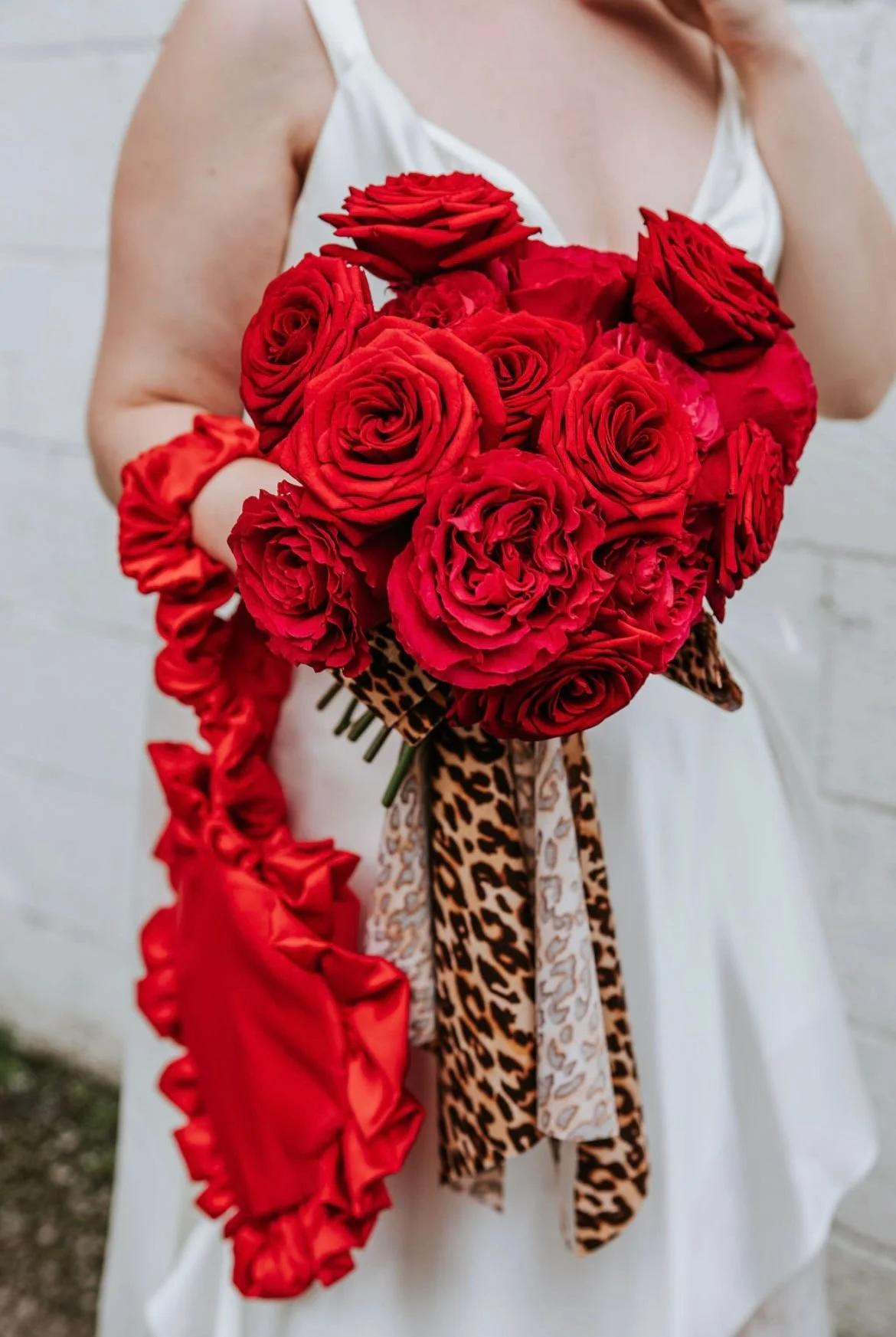 Person in a white dress holding a bouquet of red roses decorated with animal print and satin ribbons.