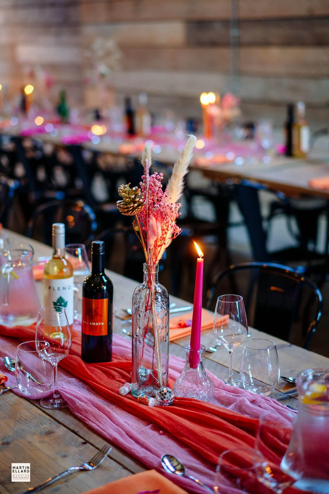 Decorated table setting with wine bottles, glasses, a pink candle, and a floral centerpiece on a pink table runner at a festive event.
