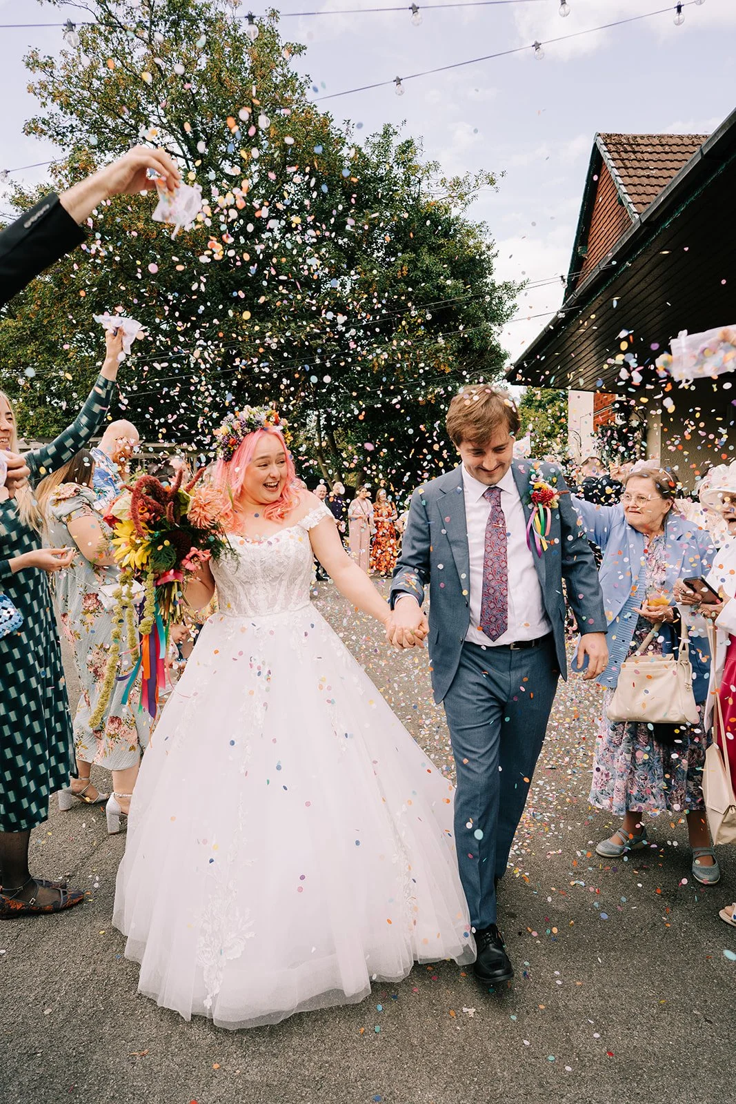 A newlywed couple holding hands and walking under colorful confetti thrown by wedding guests outdoors. The bride has pink hair and wears a white wedding dress and crown of flowers, holding a bouquet. The groom wears a blue suit with a floral tie. Guests surround them cheering.