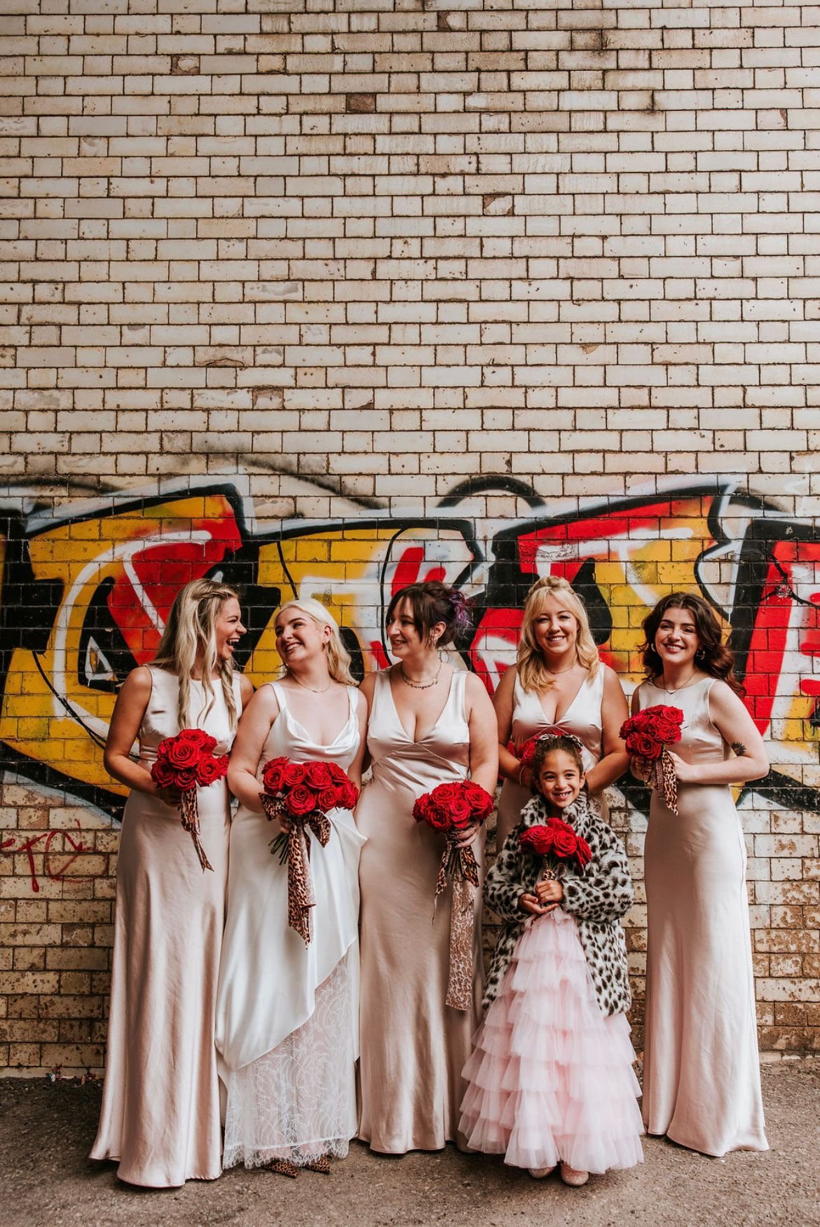 A group of six women and one young girl dressed in elegant dresses, holding bouquets of red roses, standing against a graffiti-covered brick wall. They appear to be celebrating, smiling and laughing together.