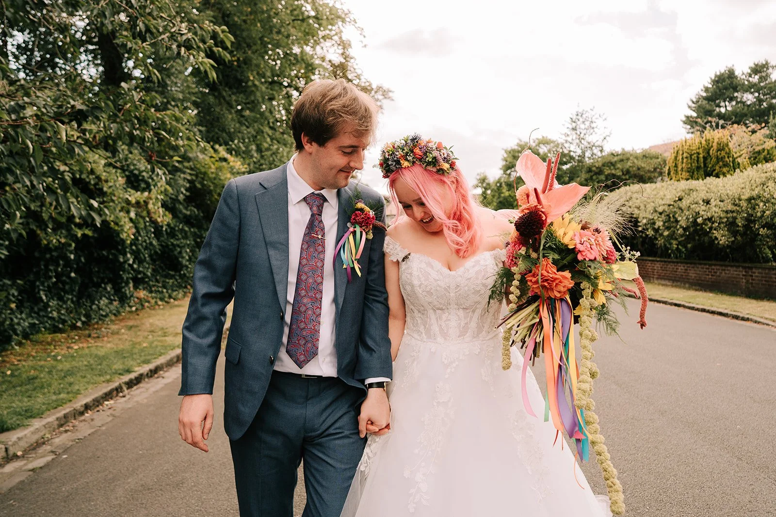 A newlywed couple holding hands and walking outdoors on a pathway, smiling and looking at each other. The woman is in a white lace wedding dress with pink hair and a floral crown, carrying a colorful bouquet. The man is in a blue suit with a patterned tie, with greenery and trees in the background.