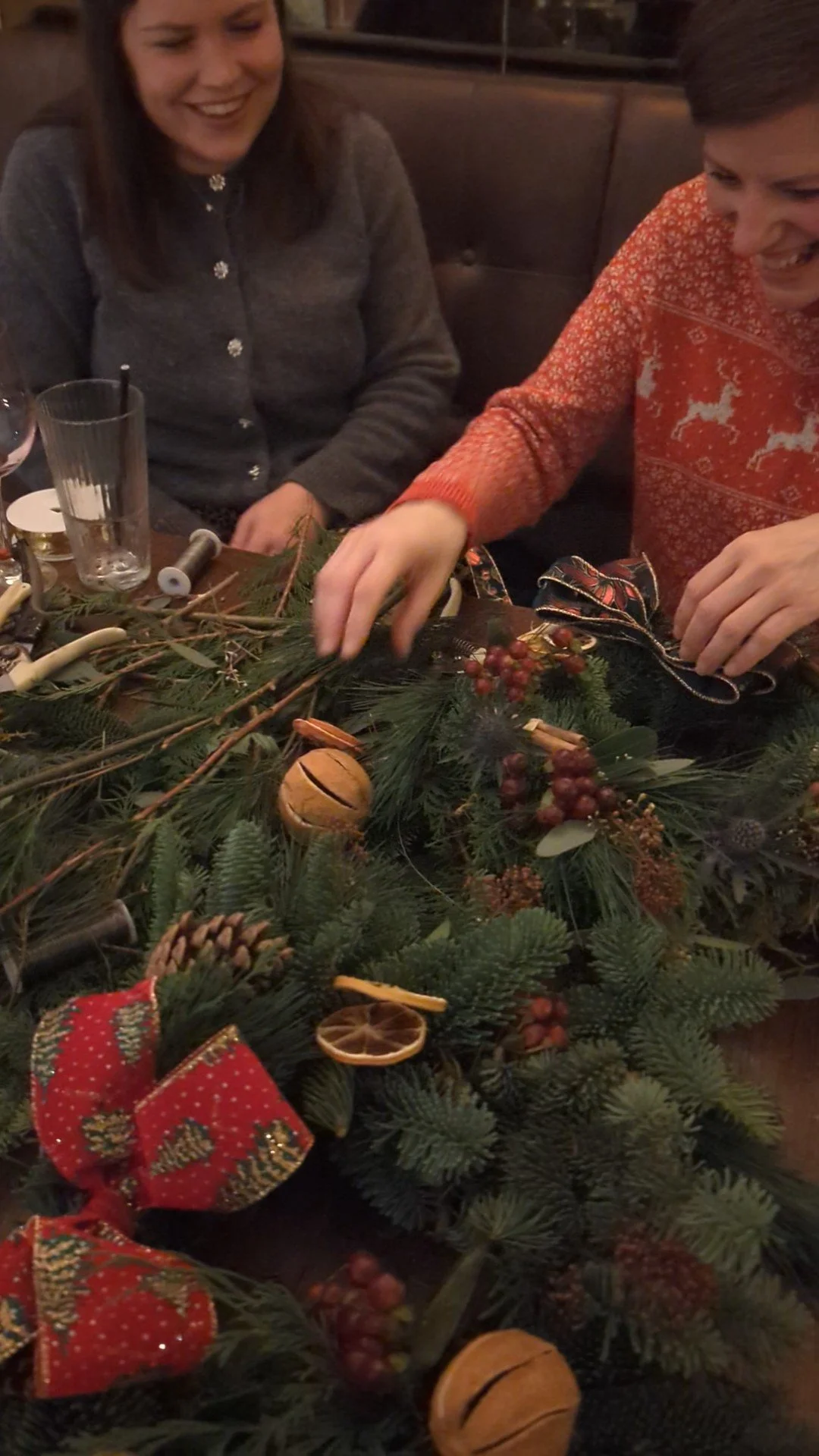 Two women sitting at a table, decorating a Christmas wreath with pine branches, red berries, dried orange slices, cinnamon sticks, and a red ribbon with Christmas patterns.