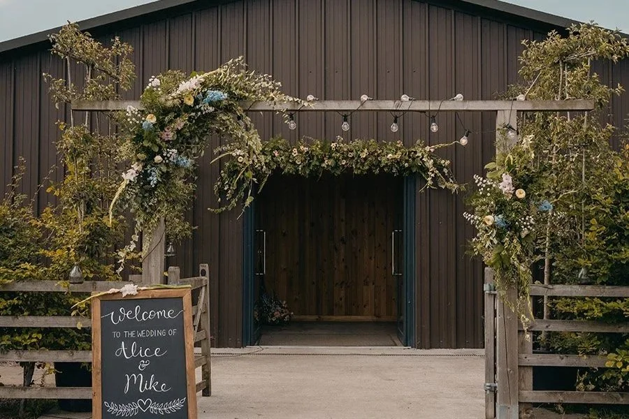 Barn decorated with floral arrangements and string lights at a wedding reception, with a welcome sign for Alice and Mike.