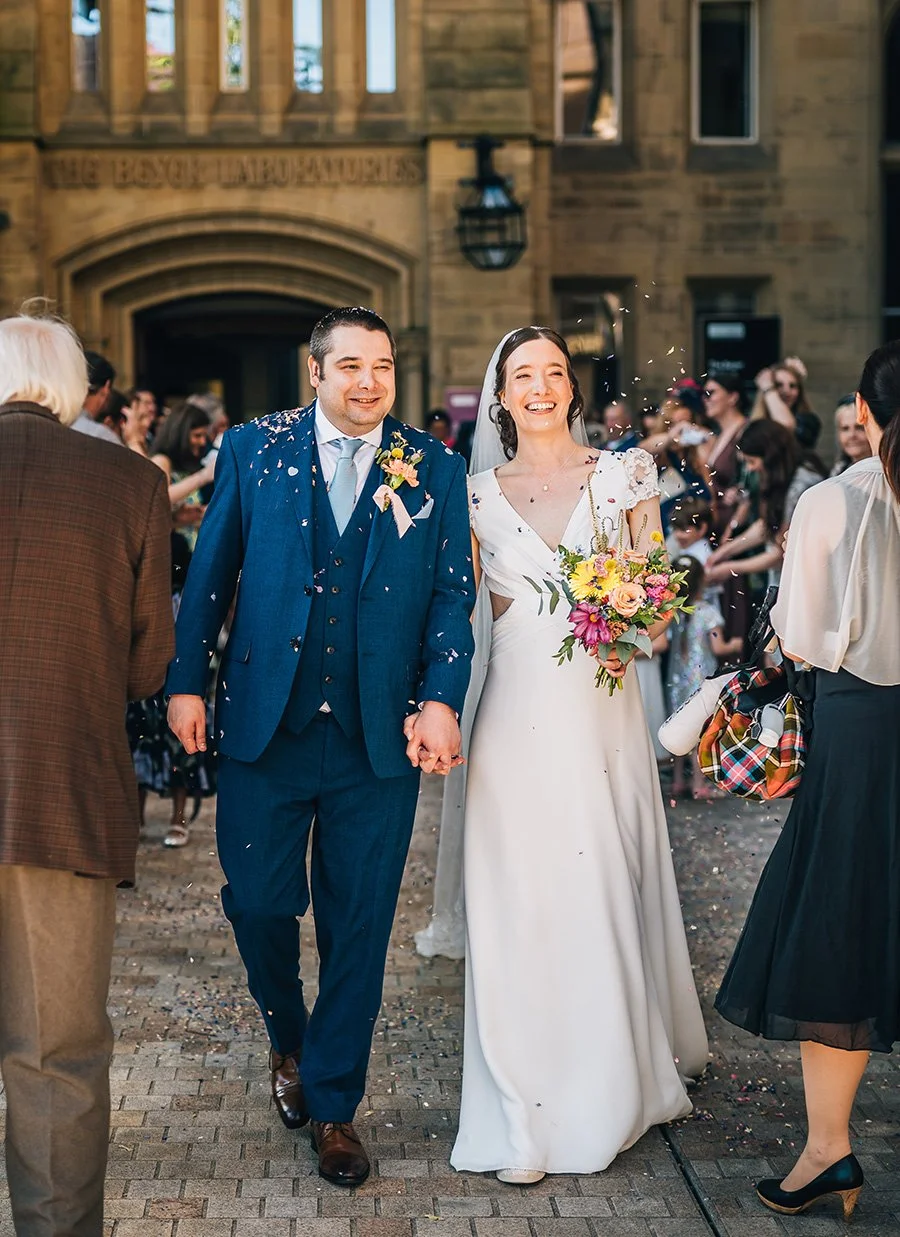 A happy bride and groom holding hands walk through a crowd of wedding guests outside a church, with confetti in the air, on a sunny day.