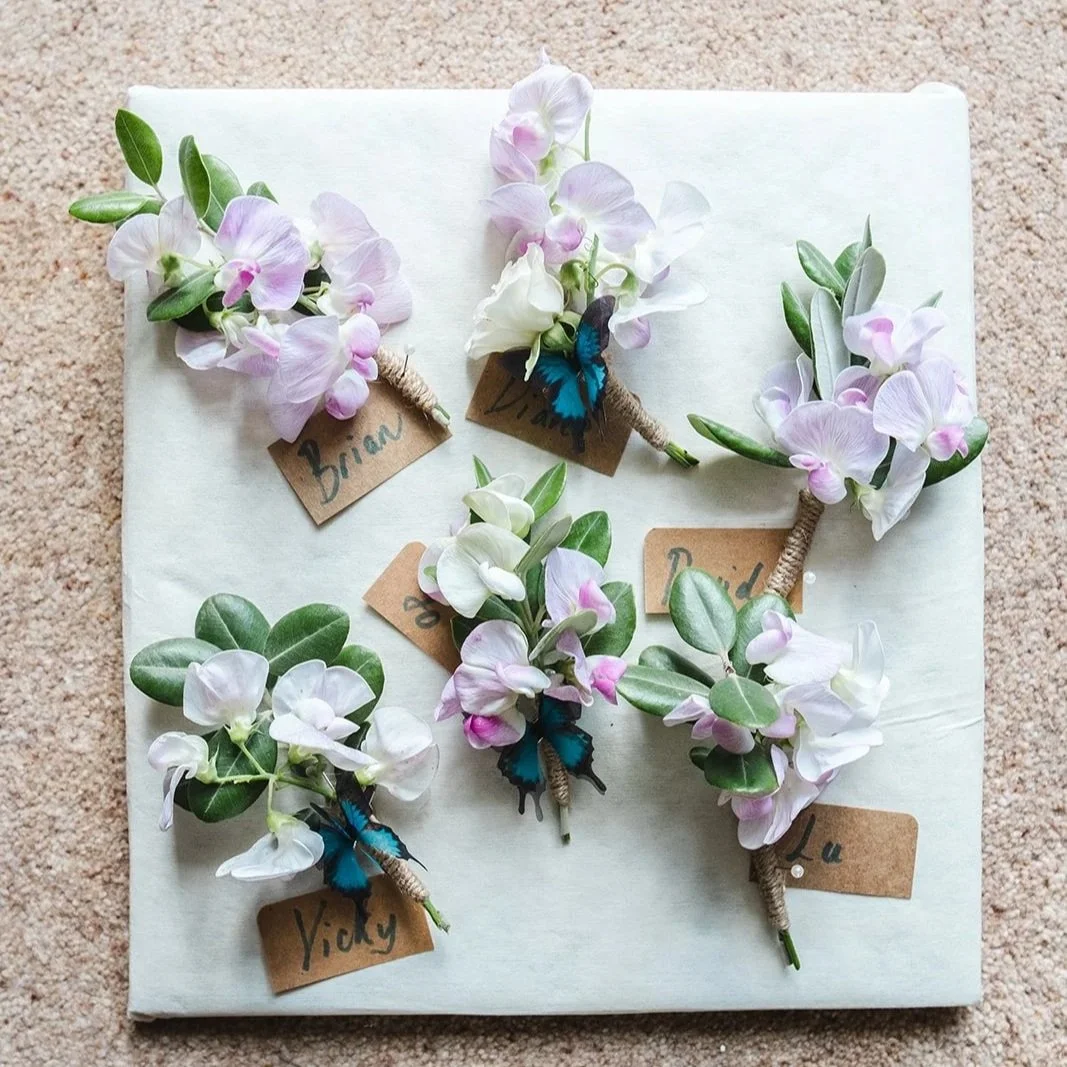 Five small boutonnières with pink and white flowers, green leaves, and blue butterflies, each labeled with a handwritten name on brown paper tags, arranged on a light-colored surface.