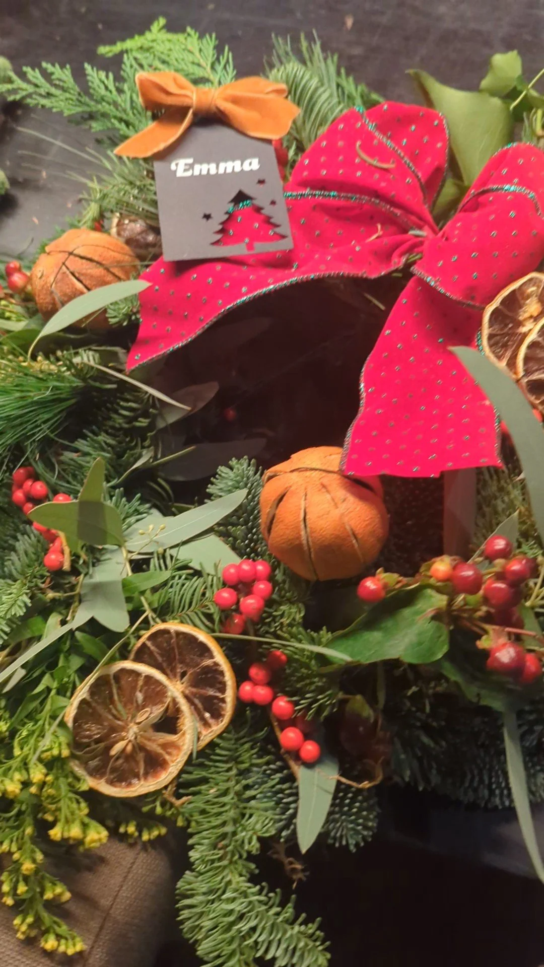 A Christmas wreath decorated with green foliage, dried orange slices, small pumpkins, red berries, a red bow with green stitching, and a gift tag with the name Emma and a cutout Christmas tree.