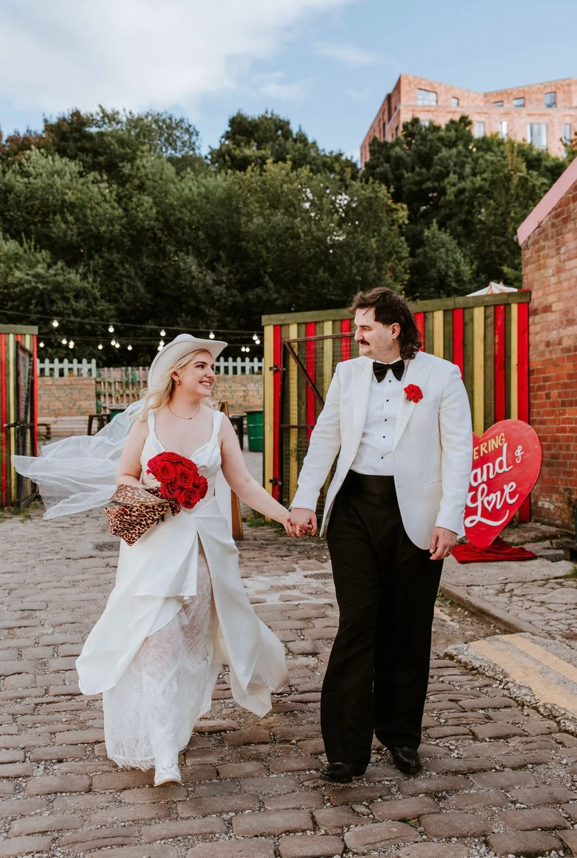 A bride and groom holding hands and walking on a cobblestone street, smiling at each other during their wedding celebration outdoors, with a colorful fence and a heart-shaped sign in the background.