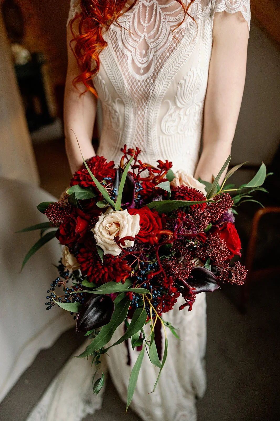 Bride in a white lace wedding dress holding a bouquet of red, white, and dark purple flowers with green leaves.