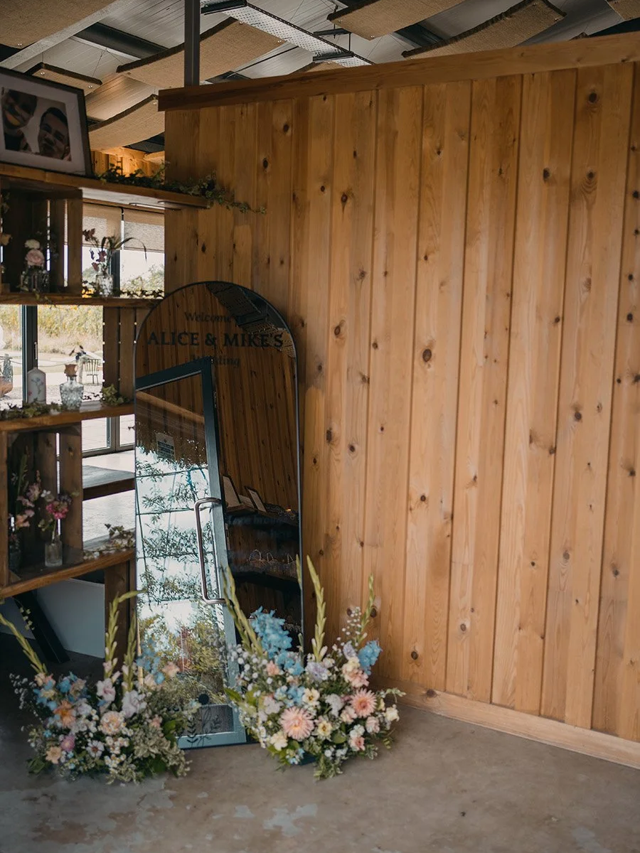 Wood-paneled wall with a floral arrangement and a mirror leaning against it, reflecting the venue entrance with a sign that says "Welcome! Alice & Mike".
