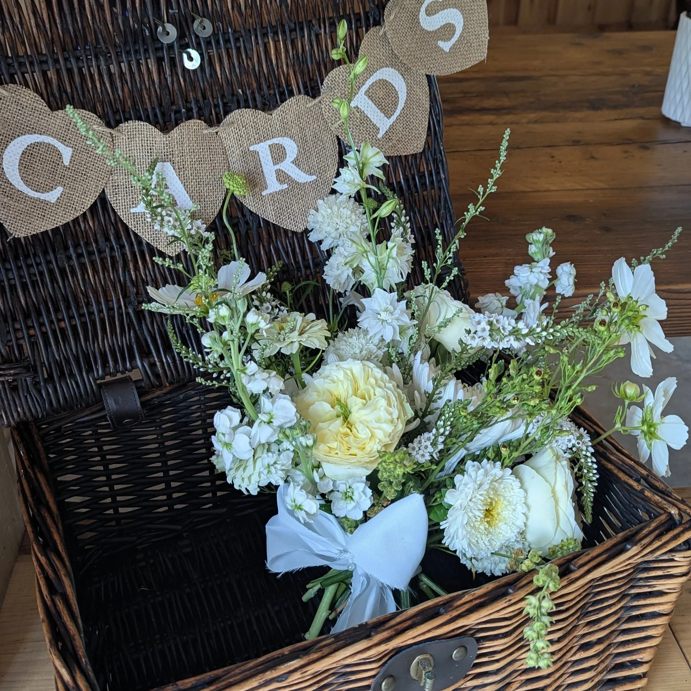 A black wicker basket with white and yellow flowers inside, placed on a wooden surface, decorated with a burlap banner with white letters spelling 'CROSS' strung across the basket.