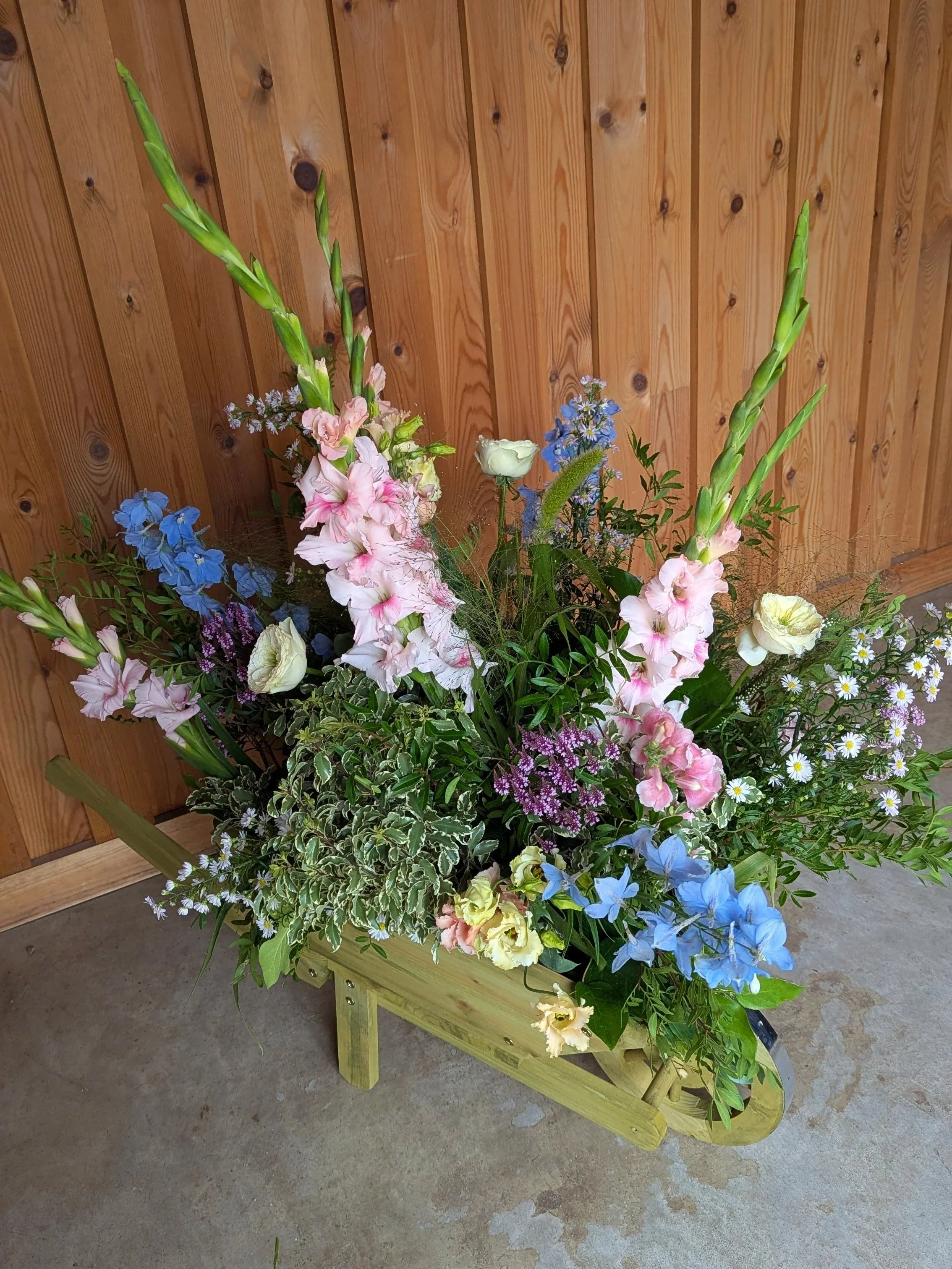A large arrangement of pink, blue, white, and purple flowers in a green wooden cart with a wooden wall background.