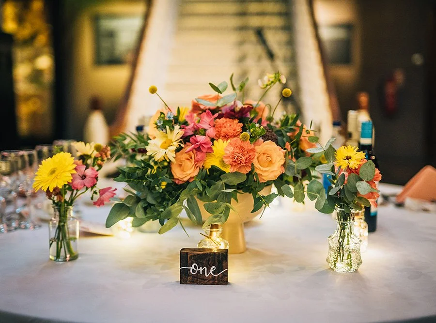 A large floral centerpiece with pink, yellow, orange flowers and green leaves on a white table. Small matching vases with flowers are on either side. A blackboard with the word "One" is in front of the centerpiece. The background shows a blurred outdoor or indoor setting, possibly at a celebration or event.