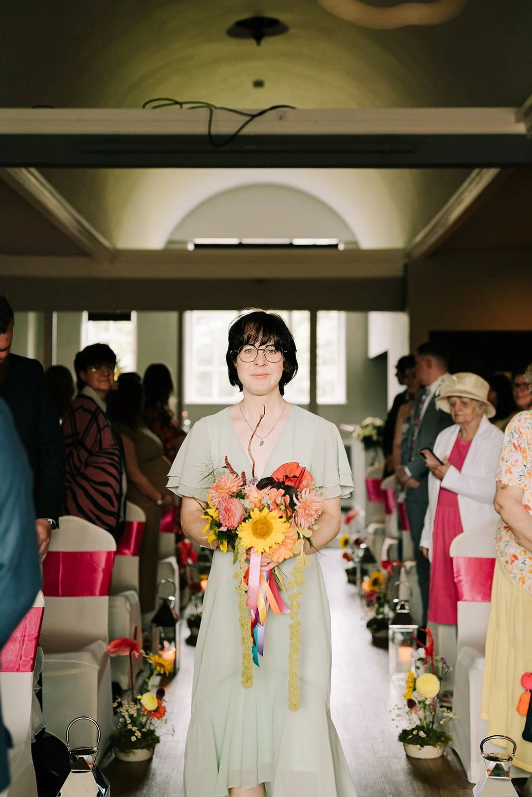 Bridesmaid with colourful bouquet walking down aisle created by MillieBuds Colour Loving Wedding Florist in Stockport at Bowdon Rooms