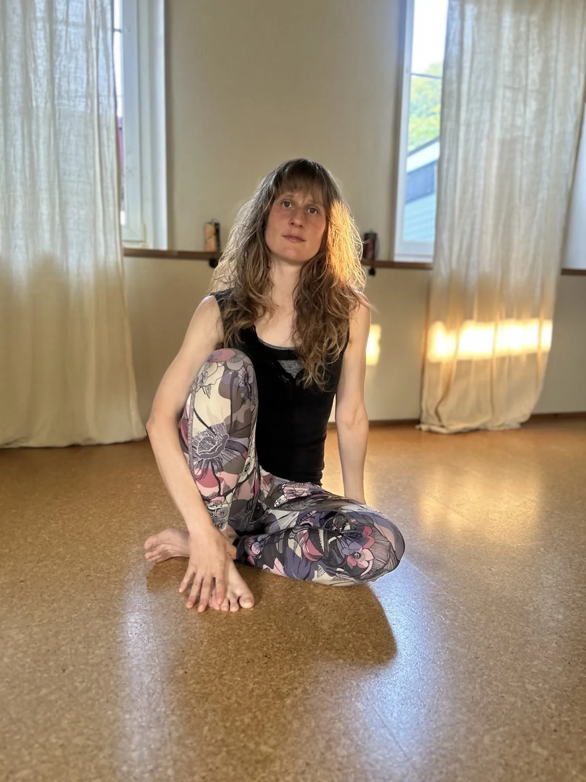 Eine Frau mit langen lockigen Haaren sitzt im Kursraum auf dem Boden, gekleidet in einem schwarzen Top und bunten, floral bedruckten Leggings, umgeben von Vorhängen und natürlichem Licht.
