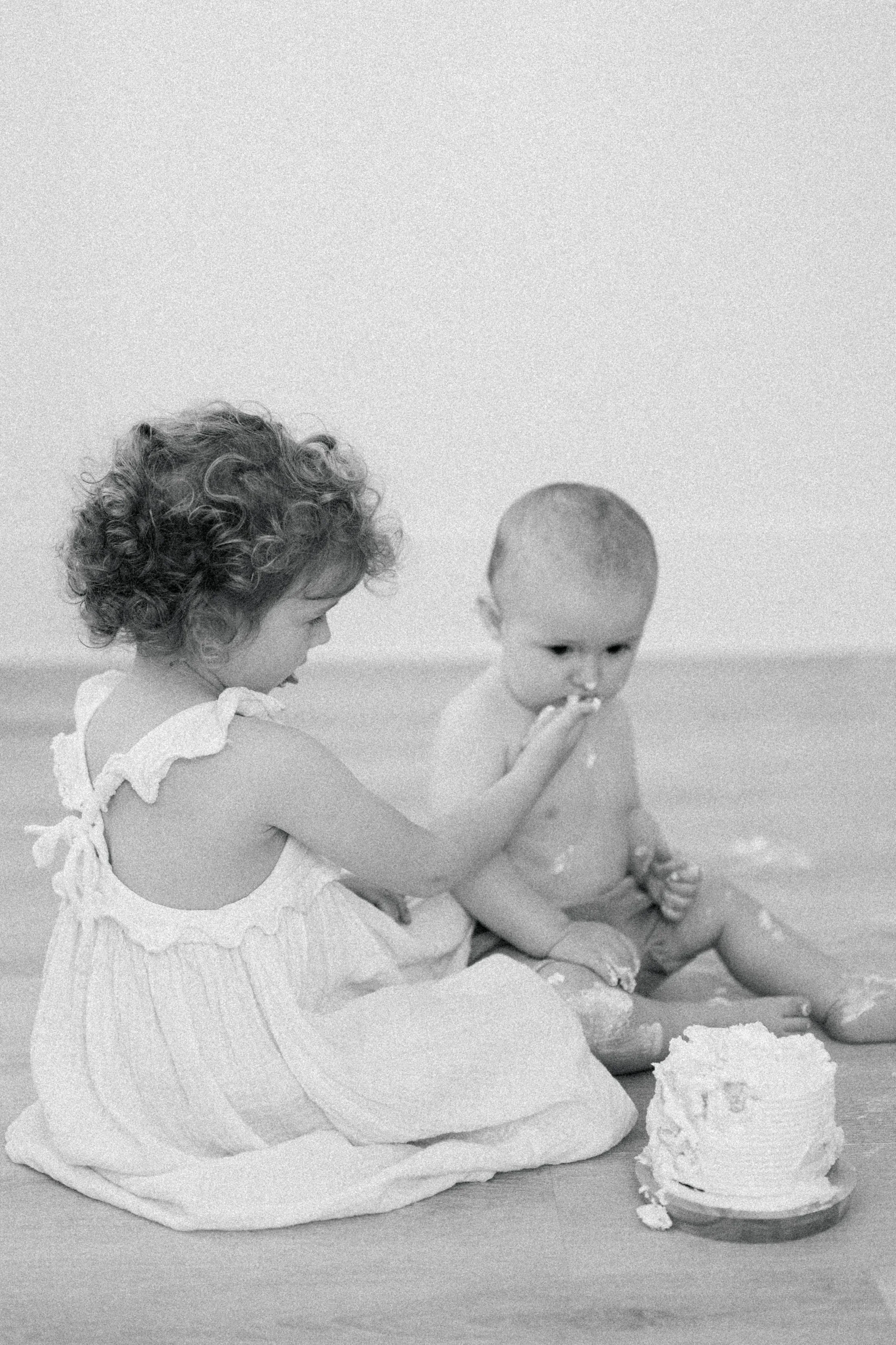 A young girl and a baby sitting on a floor with a cake in front of them. The girl is feeding the baby with her finger.