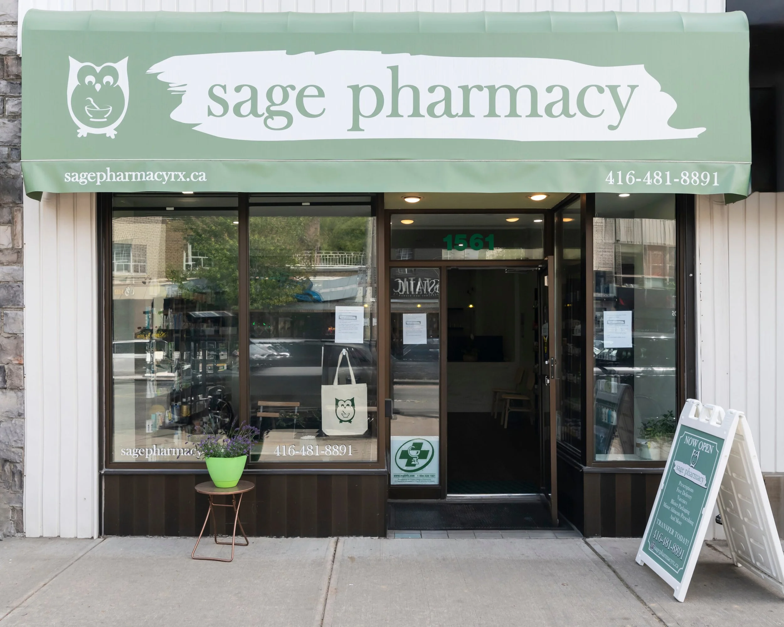 Front entrance of Sage Pharmacy with a green awning displaying the pharmacy's name, logo with an owl, website, and phone number. A small side table with a green potted plant and a chalkboard sign outside the store.