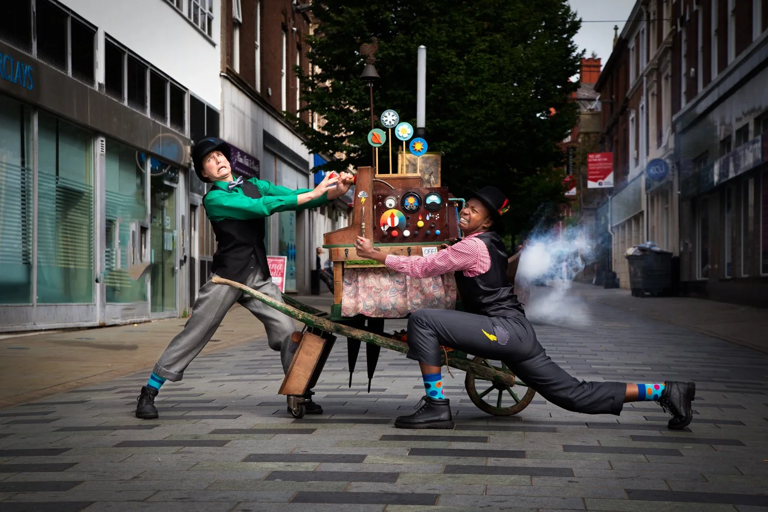 Two people in suits pushing a steam machine in a high street. They look to be struggling with the machine. 