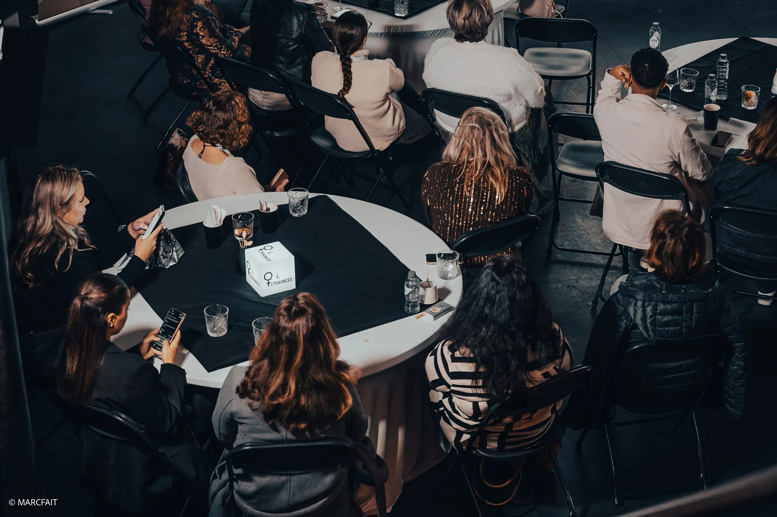 Group of people seated around a round conference table during a business event or conference, some using smartphones, with bottled water and glasses on the table.