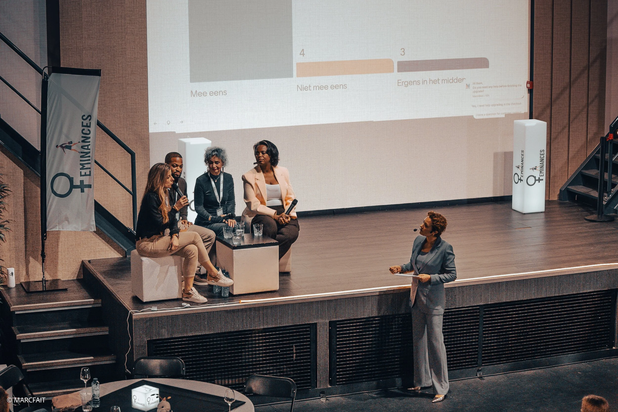 A panel discussion at a conference with four women seated on a stage, one woman standing with a microphone, and a large screen behind displaying a chart with text in Dutch. The conference branding 'EMINANCES' and a symbol of female empowerment is vis