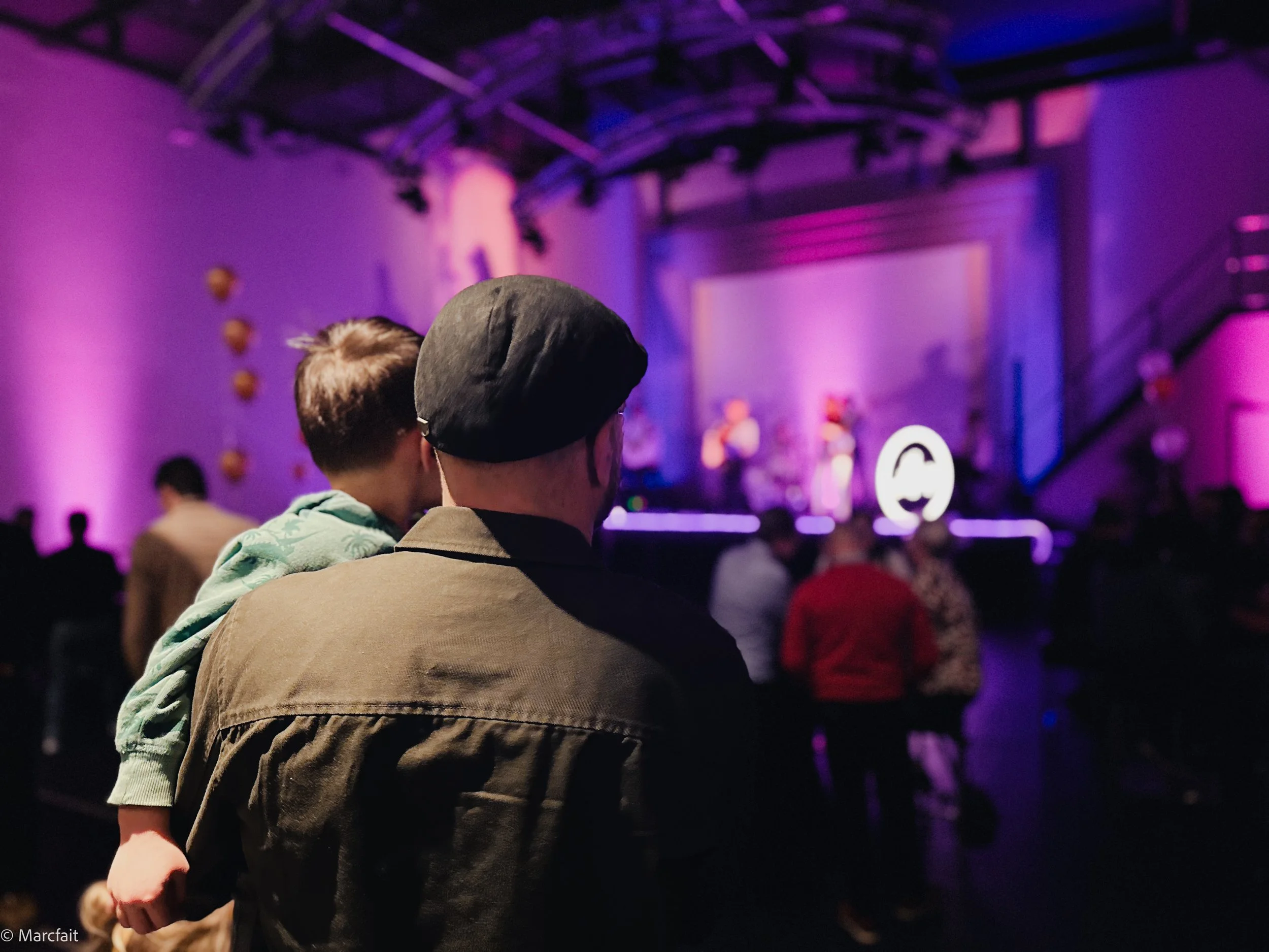 A person wearing a black beret and a brown jacket is holding a young child in a green jacket at an indoor concert or event with a stage illuminated in purple and blue lights. Other attendees are visible in the background, watching the performers.