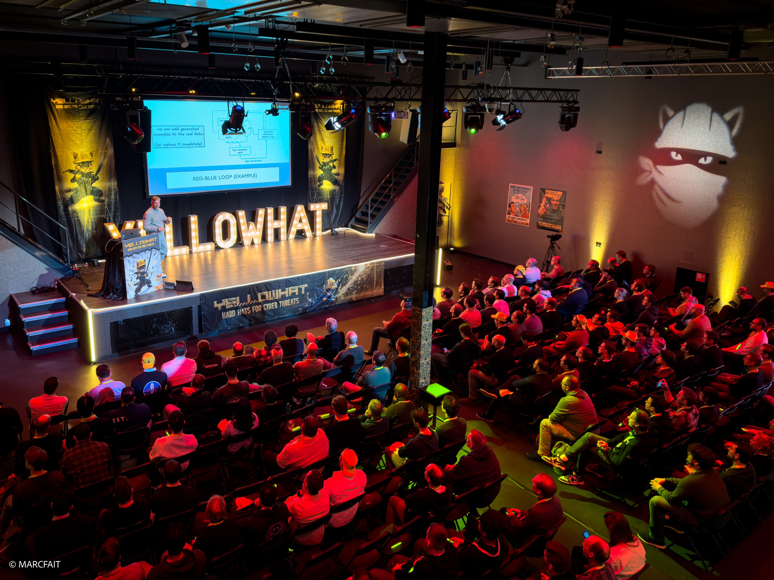 Audience watching a presentation on stage at 'YELLOWHAT', an international cybersecurity conference. The FAIT hall is decorated with posters, a large illuminated logo of a masked ninja cat on the wall, and lighting effects.