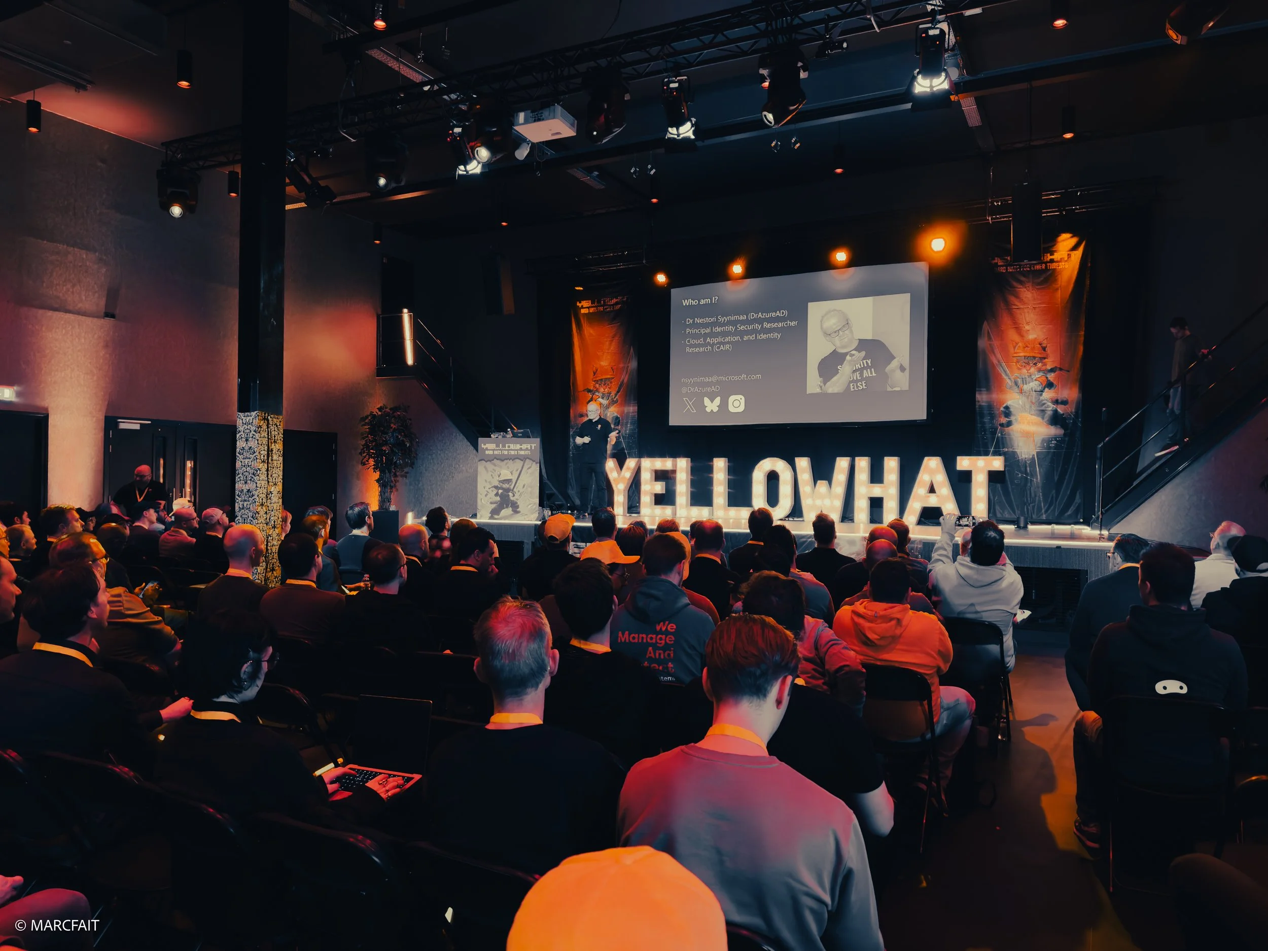 A large audience attending a conference or presentation in a darkened concert hall or auditorium. A speaker stands on stage in front of a projection screen with an image and text, and large illuminated letters spelling 'YELLOWHAT' are displayed behin