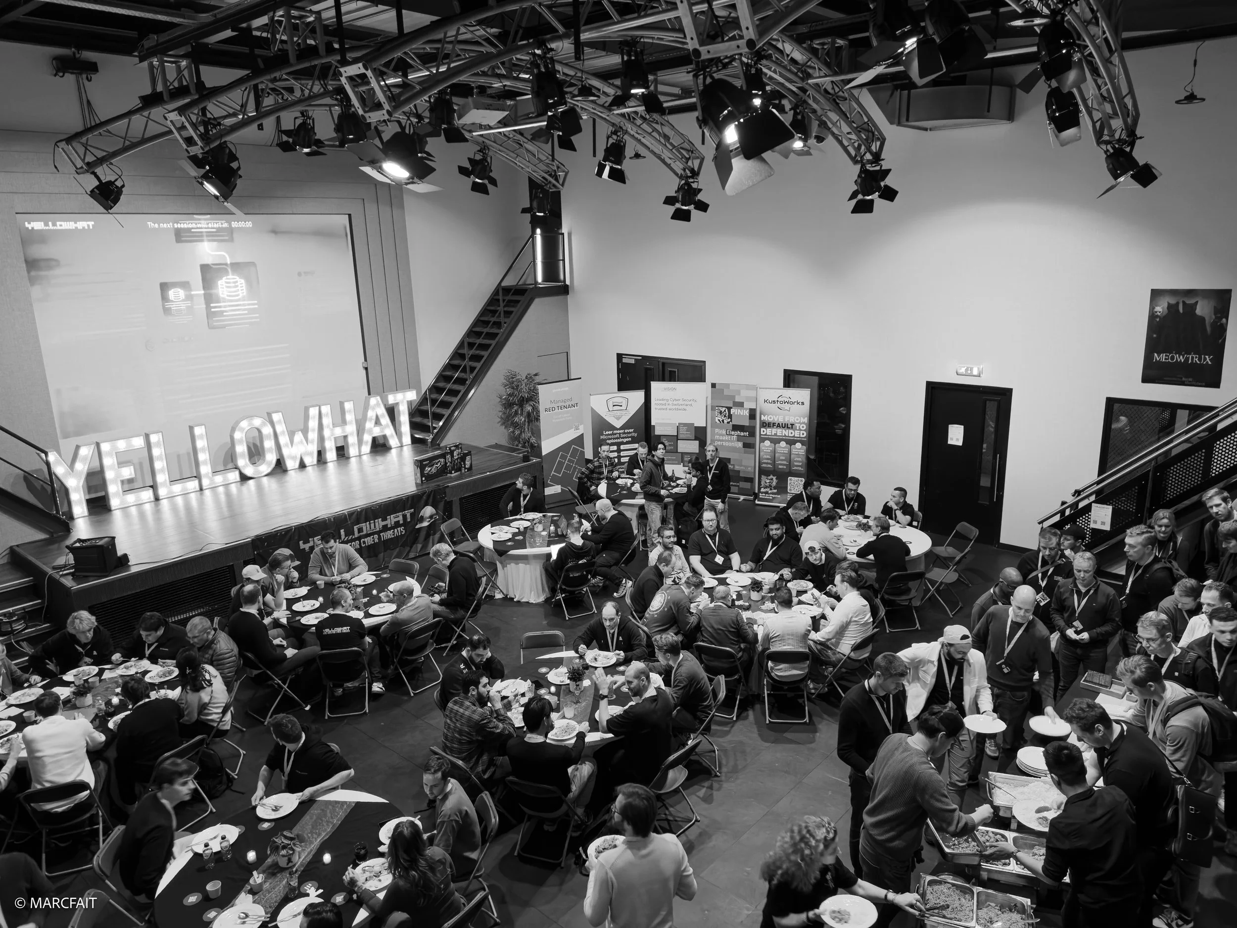 Conference or event with a large group of people seated at round tables and some standing, in a room with stage, lighting equipment, and a large screen, with signs reading "YELLOWHAT" and banners for vendors or presentations.