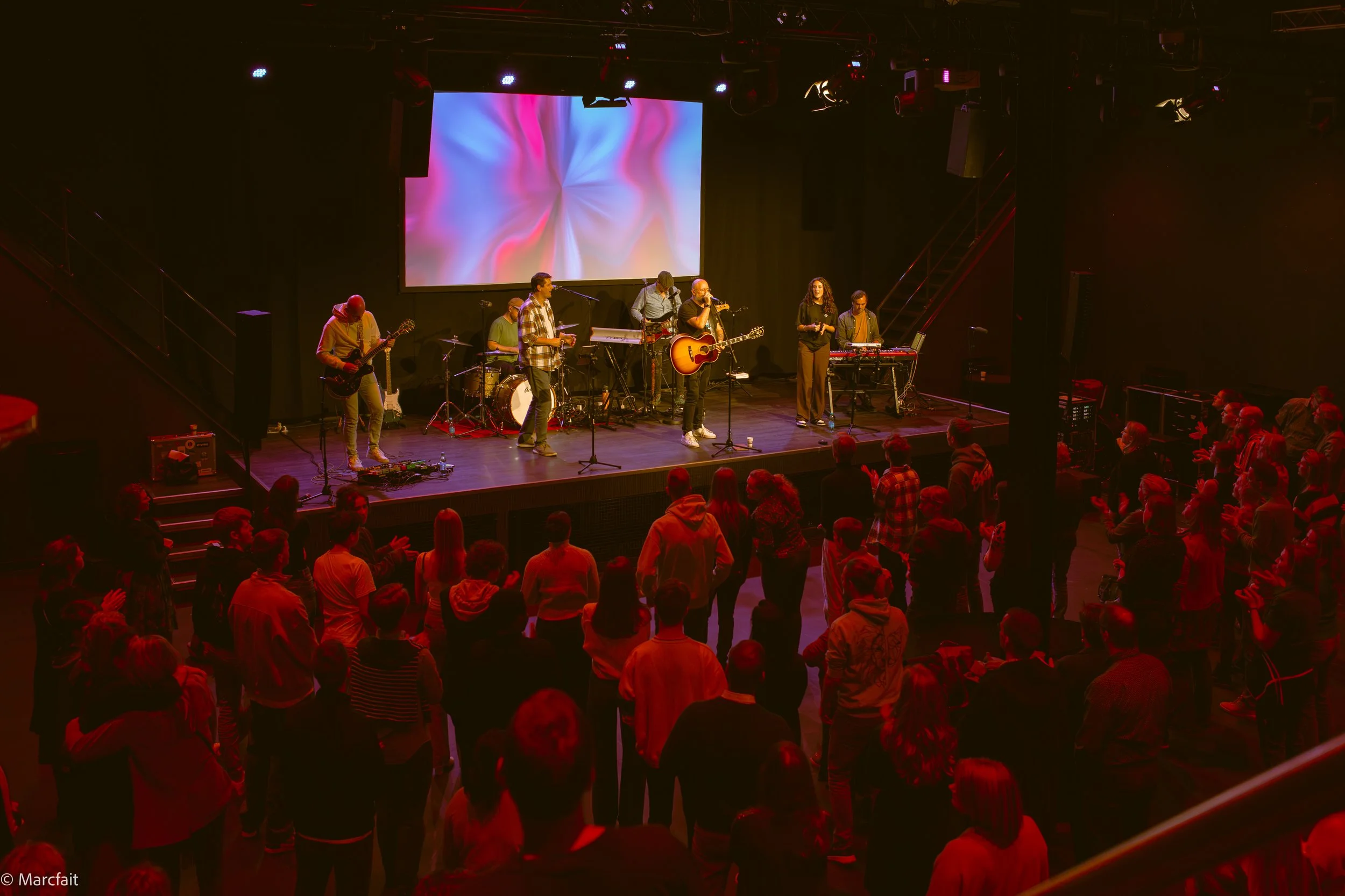 A band of seven musicians performing on stage with screens and lights overhead, audience watching and clapping in front of the stage in a dimly lit concert venue.