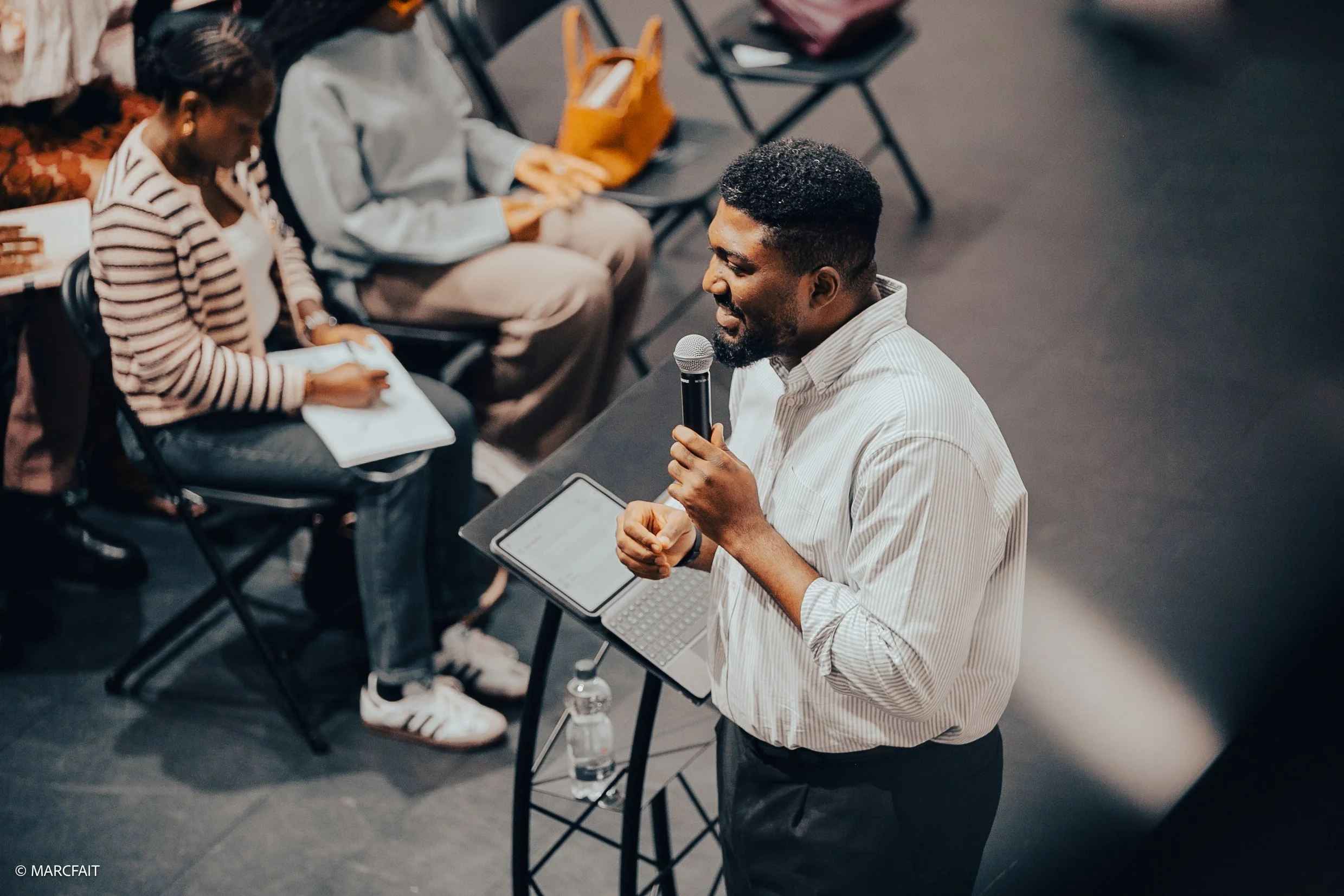 Man giving a presentation with a microphone, standing next to a table with a laptop, in front of seated audience members taking notes.