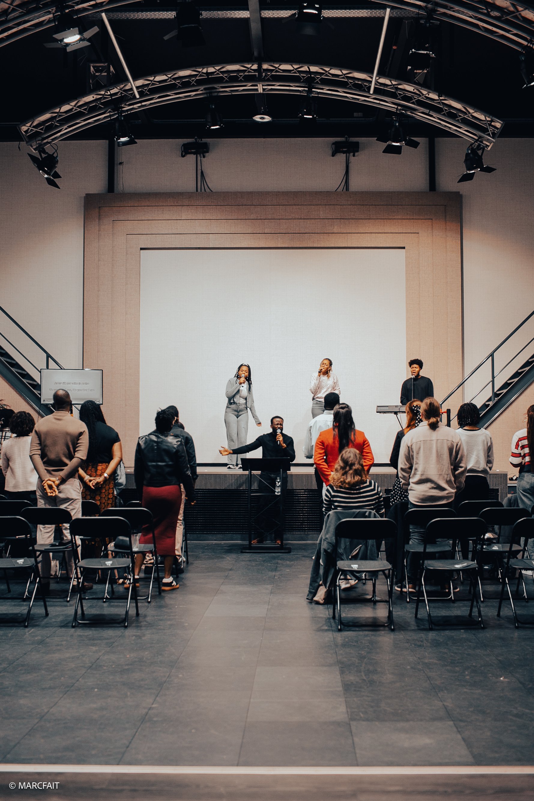A group of people is gathered in a large auditorium, participating in a live performance or presentation on a stage with four singers and a keyboard player. Everyone is standing, facing the stage, with some engaged and others observing.