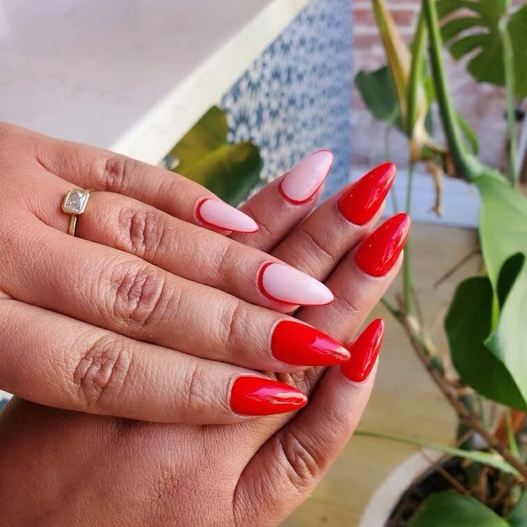 Close-up of a person's hands with manicured nails painted bright red and light pink with red outlines, wearing a silver ring on the ring finger of the left hand, with green plants and a brick wall in the background.