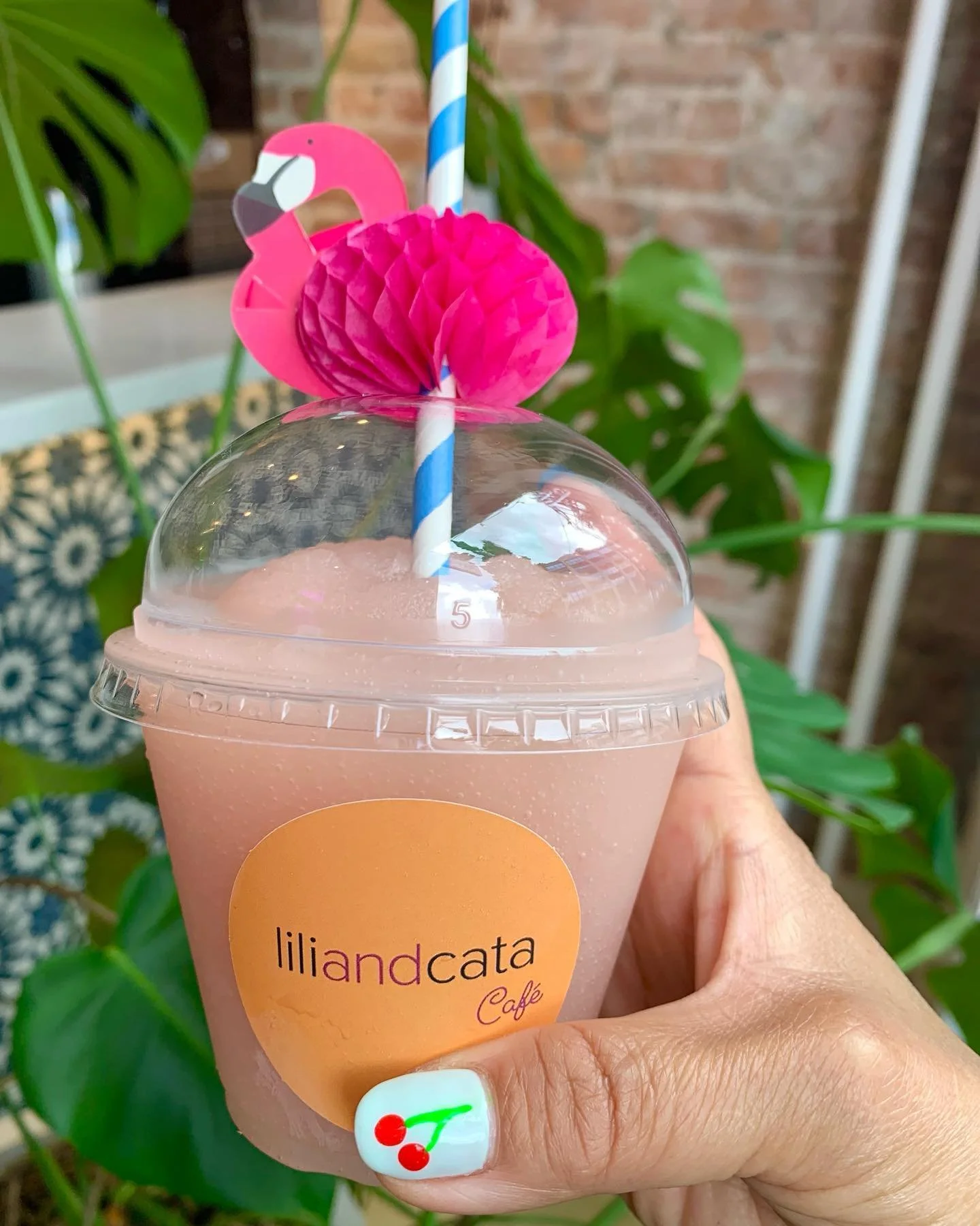 Hand holding a clear plastic cup with frozé in it, decorated with a pink flamingo paper straw, surrounded with green plants and a brick wall in the background.