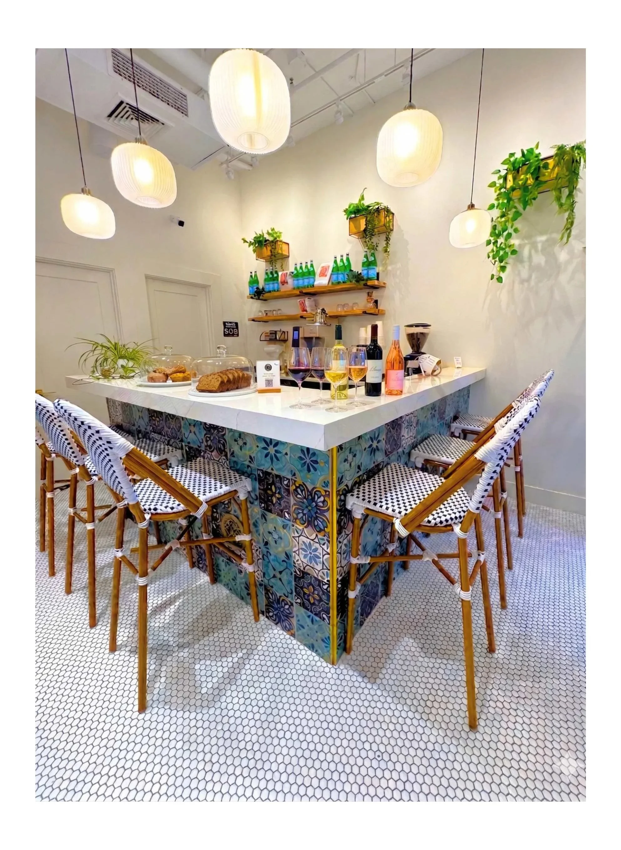 A stylish indoor bar area with a white countertop, decorated with patterned tiles, surrounded by bar stools with black and white patterned seats. The bar has bottles of wine and water, glasses of red and white wine, and baked goods under glass covers. The background features shelves with bottles, plants, and glassware, and is illuminated by hanging white pendant lights.