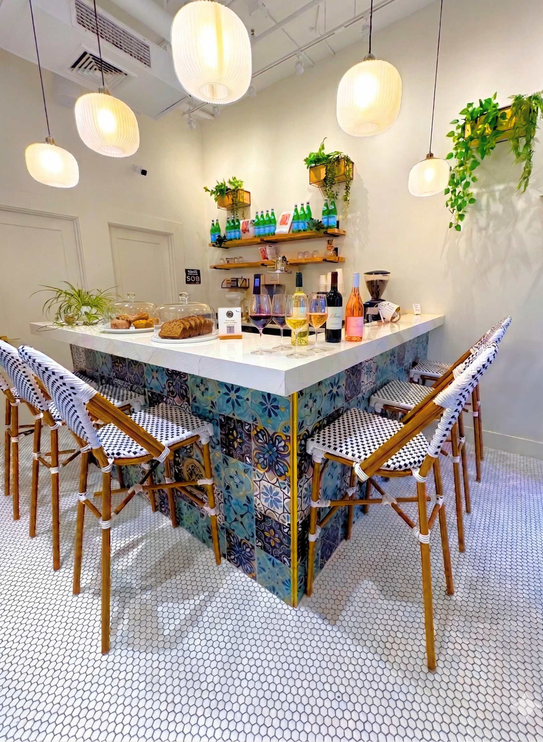 A stylish dining space with a corner bar surrounded by four chairs with woven black and white seats. The bar has a white tile counter with colorful patterned tiles on the lower side. There are various bottles and wine glasses on the counter, along with a cake under a glass dome. Hanging above are six white paper lantern pendant lights, and the background features white walls, green plants, and wooden shelves with more bottles and glasses.