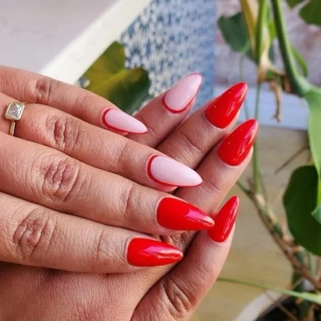 Close-up of hands with red and white manicured nails, one hand with a gold ring, background of green leaves and a wooden surface.