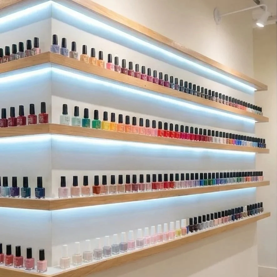 Display of multiple rows of colorful nail polish bottles on wooden shelves with LED lighting in a nail salon.