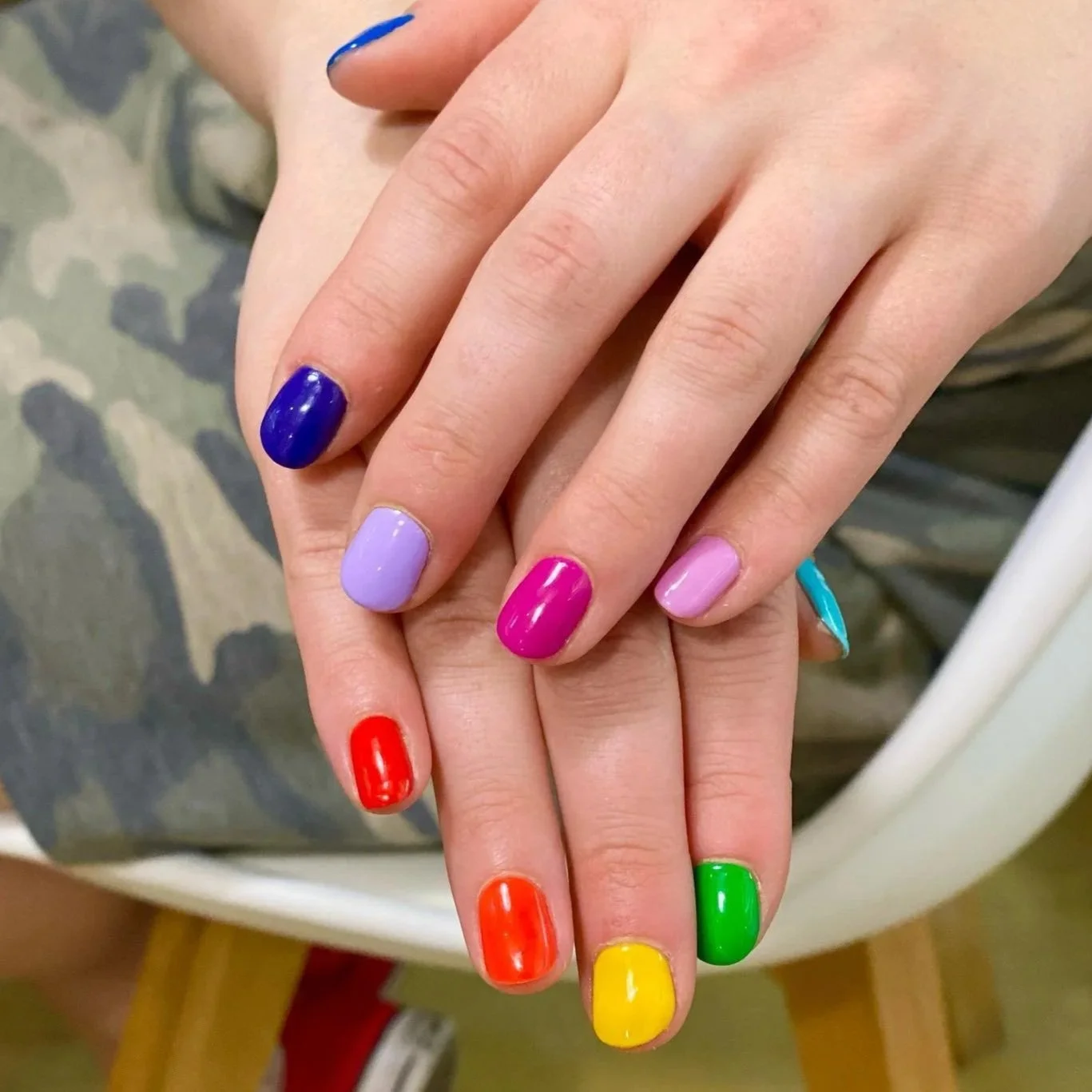 Close-up of two hands with colorful painted nails, overlapping. The nails are painted in shades of purple, pink, blue, red, green, and yellow.
