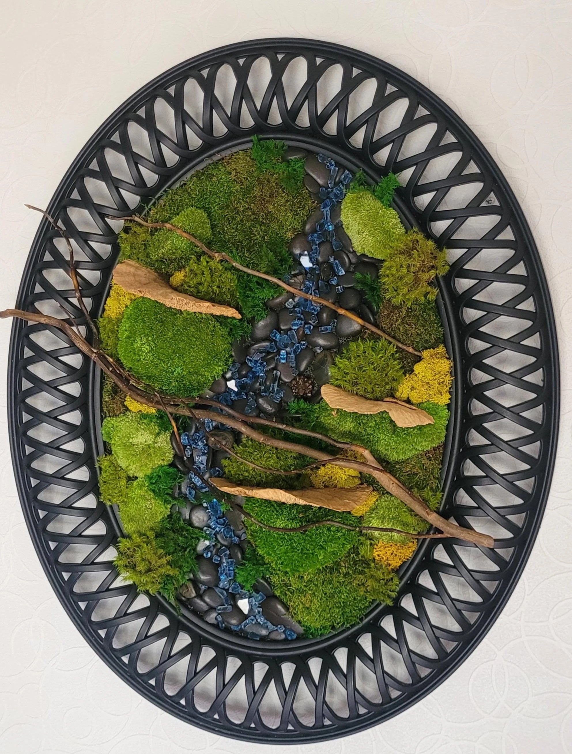 A decorative wall art piece featuring a circular black frame with an arrangement of artificial moss, twigs, stones, and blue glass pieces, resembling a miniature landscape.