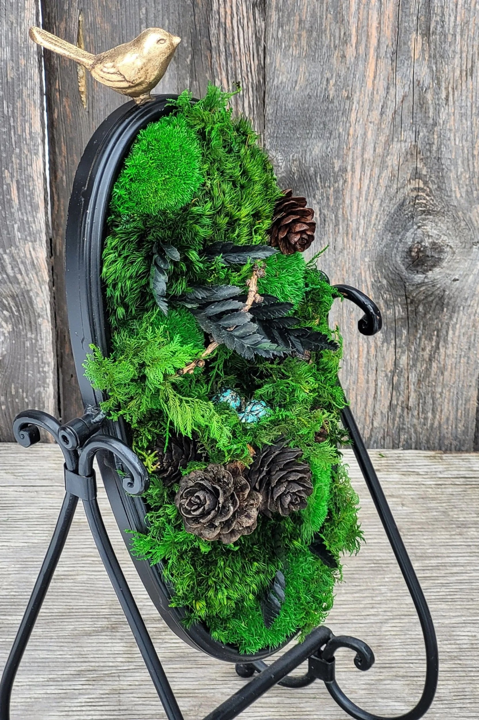 Decorative moss-covered frame on stand with pinecones and bird ornament
