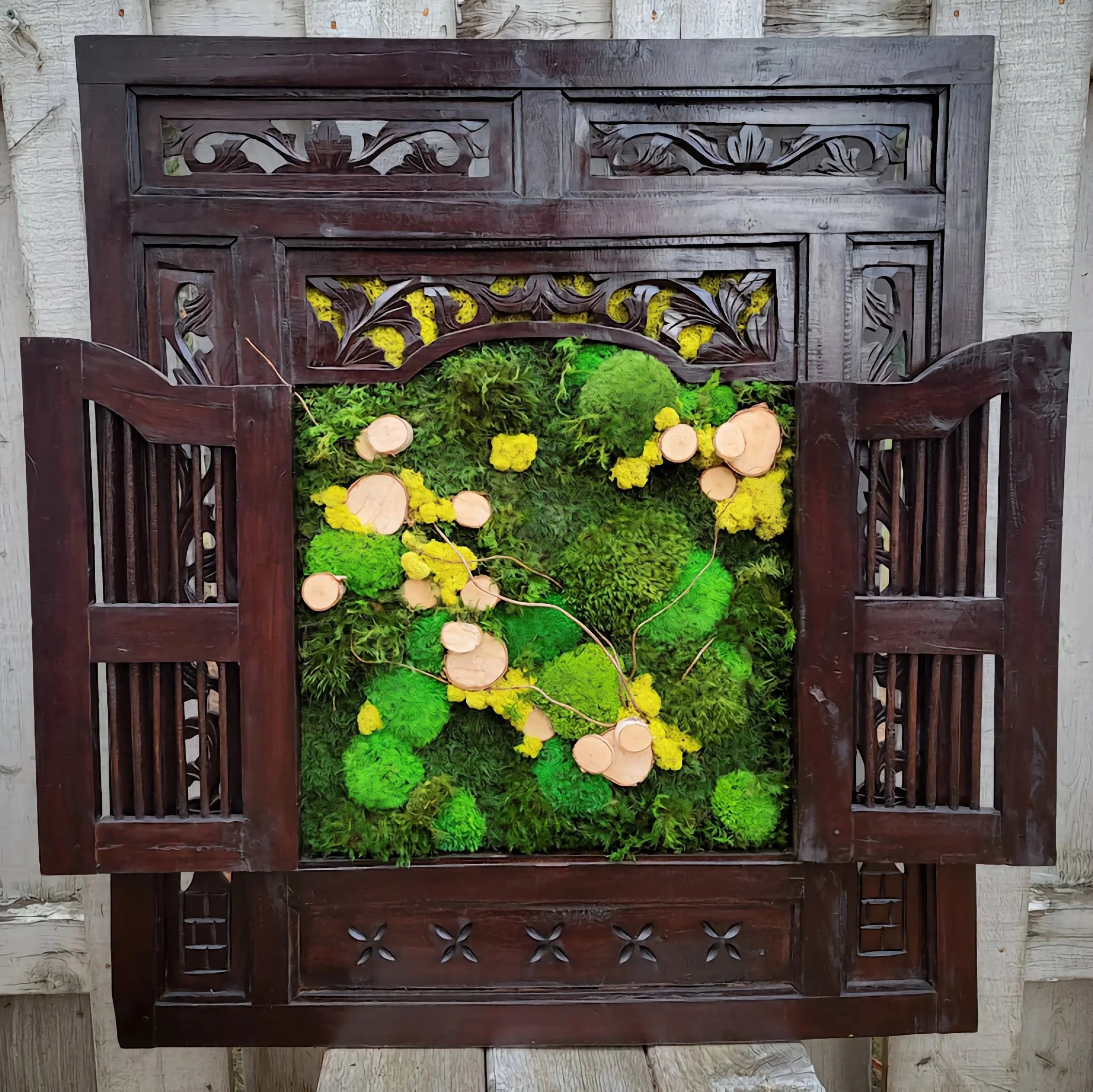 Decorative wooden frame with open shutters featuring a vertical garden of green moss, yellow flowers, and small wood slices inside.