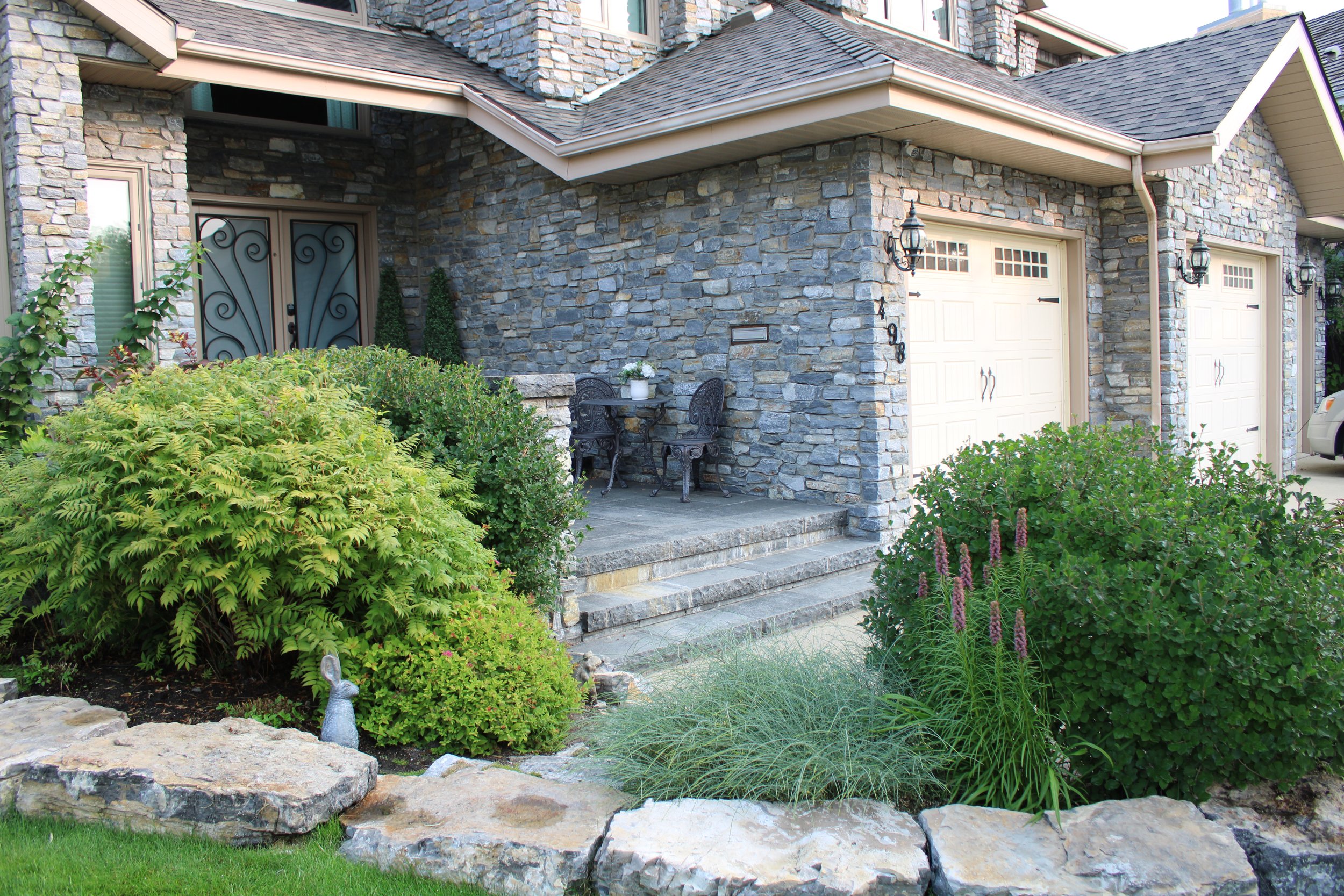 Stone house exterior with landscaped front yard, featuring bushes and ornamental grass, beside a double garage. A small patio with wrought iron chairs and table is near the entrance.