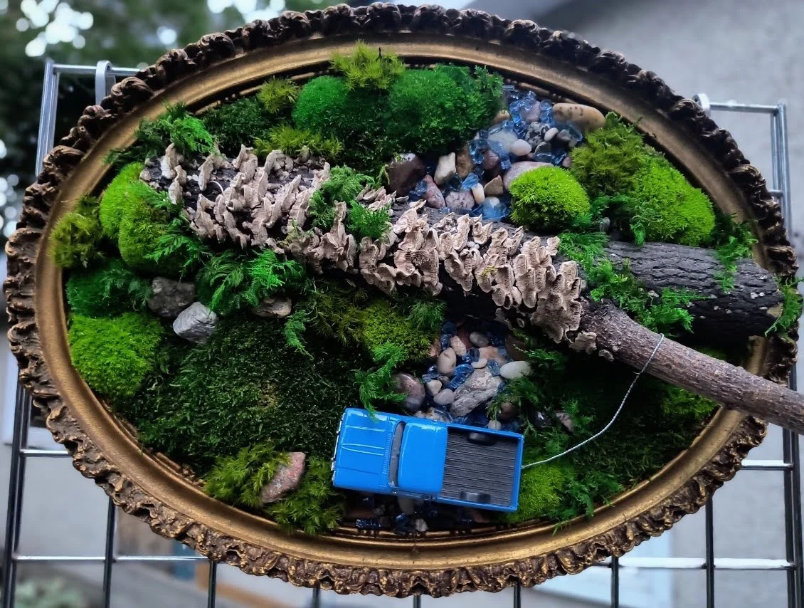 Miniature forest diorama with blue toy truck, moss, rocks, and a branch on a gold tray.