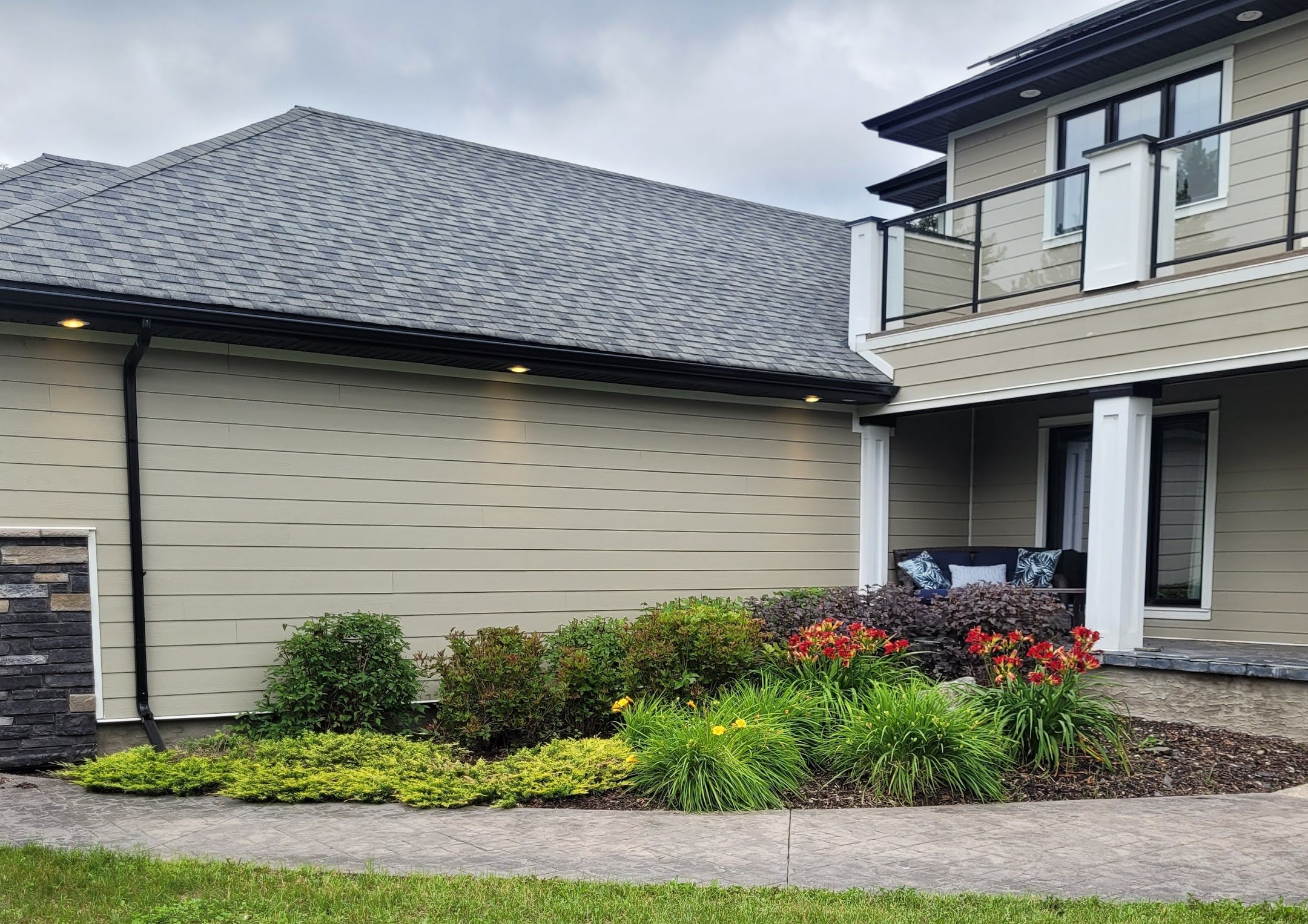Modern house exterior with beige siding, gray roof shingles, landscaped garden, stone pathway, and a balcony with glass railing.