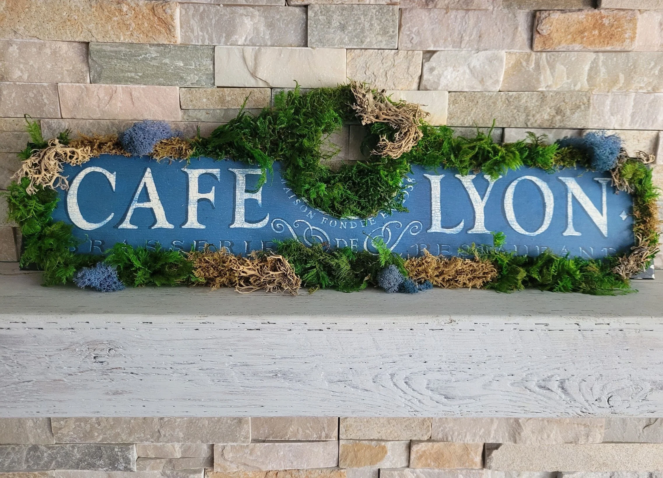 Decorative sign reading 'Cafe Lyon' on a brick wall, adorned with green moss and blue accents.